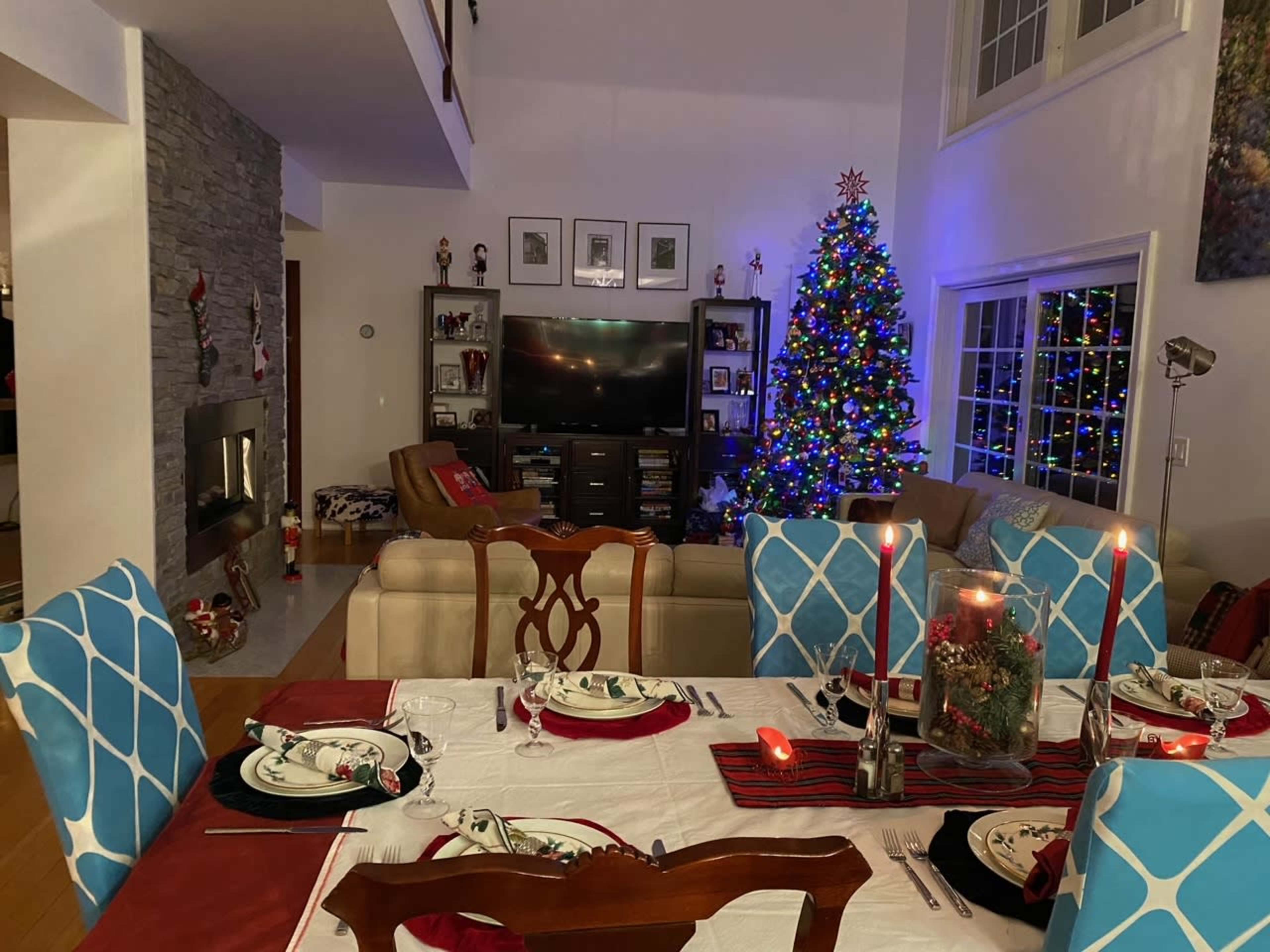 A dining table is set with plates and candles in front of a decorated Christmas tree and a living room with a large television.