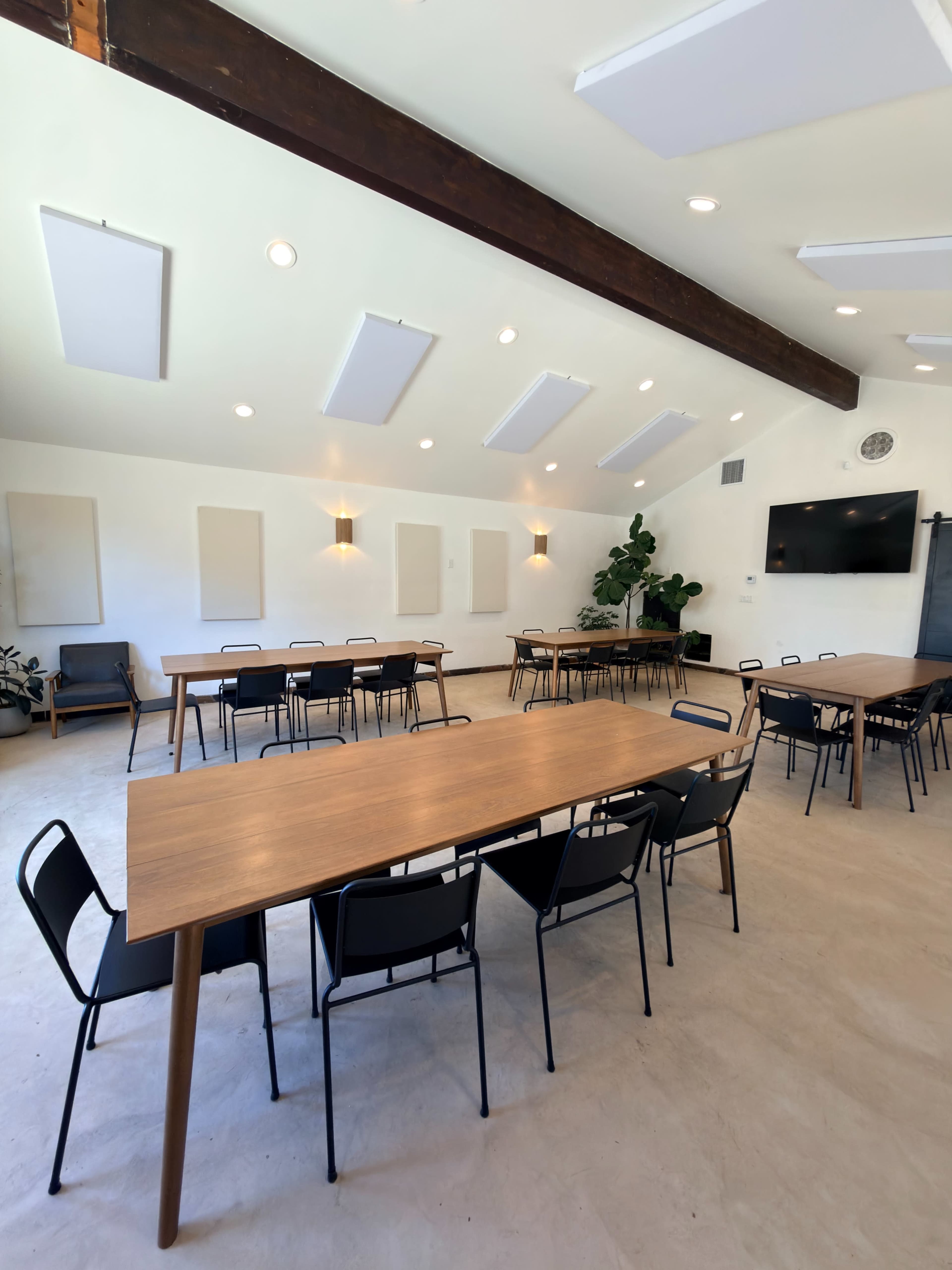 The image shows a spacious meeting room with multiple wooden tables and black chairs arranged neatly, featuring large windows and a TV screen on one wall.