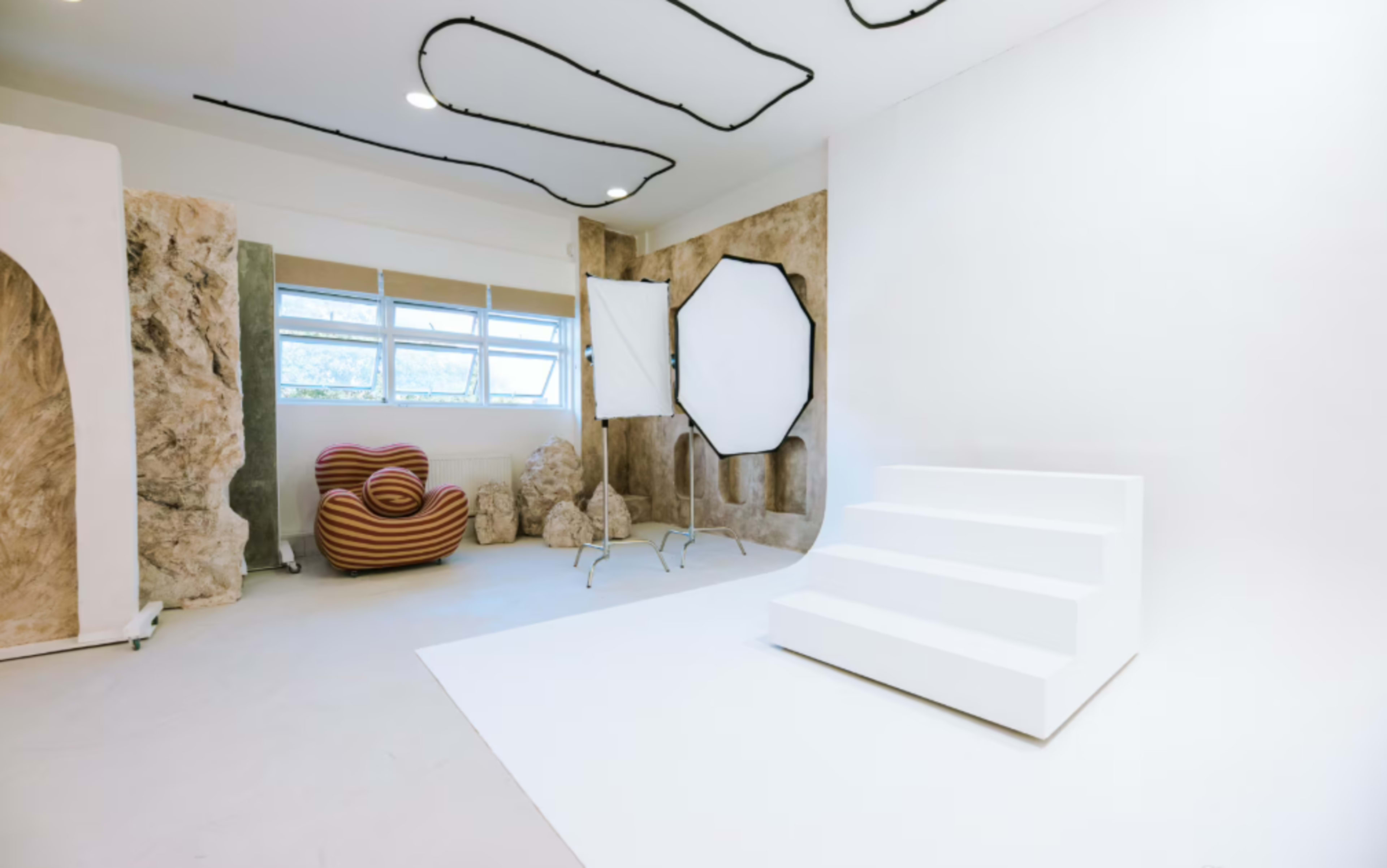 The image shows a modern photography studio with a white backdrop, three white steps, and abstract stone-like structures on the walls.