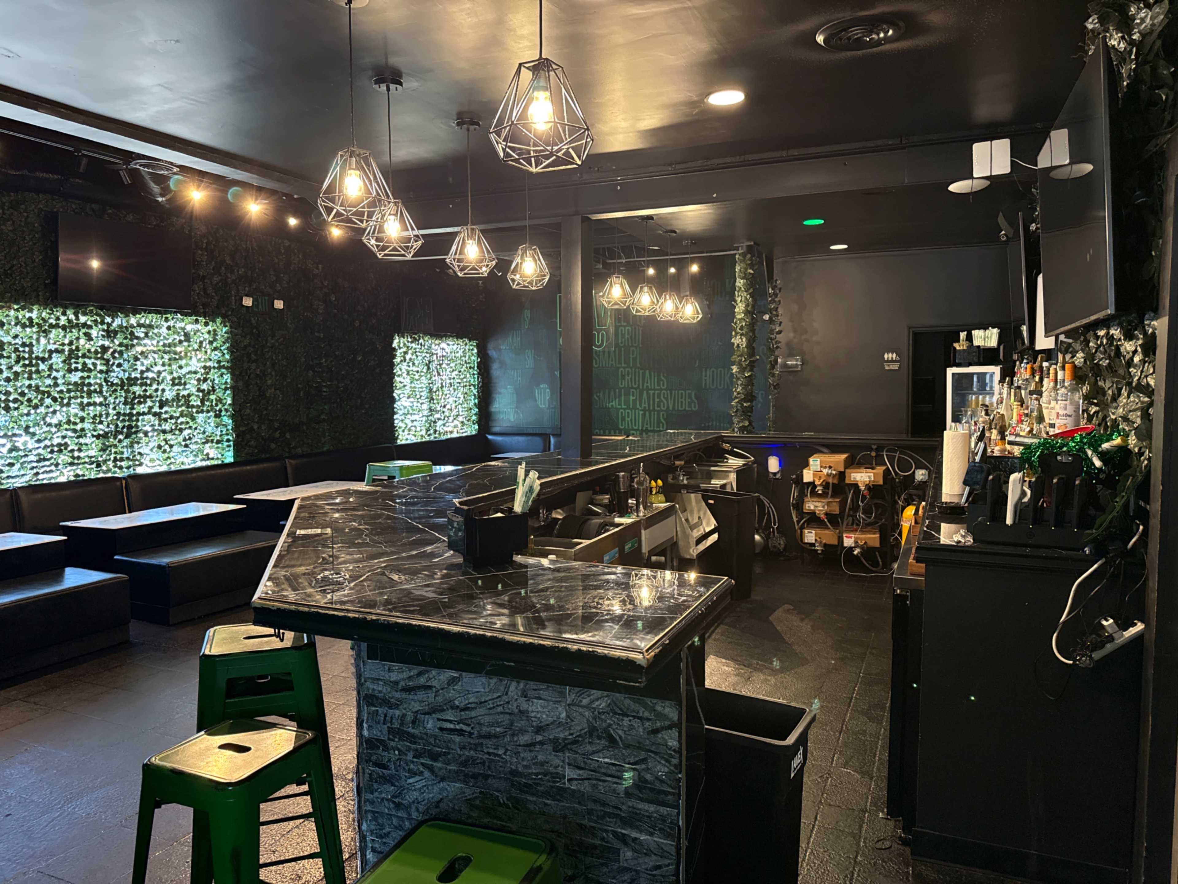 The image shows a modern bar with a marble countertop, green seating, and decorative greenery on the walls, illuminated by pendant lights.