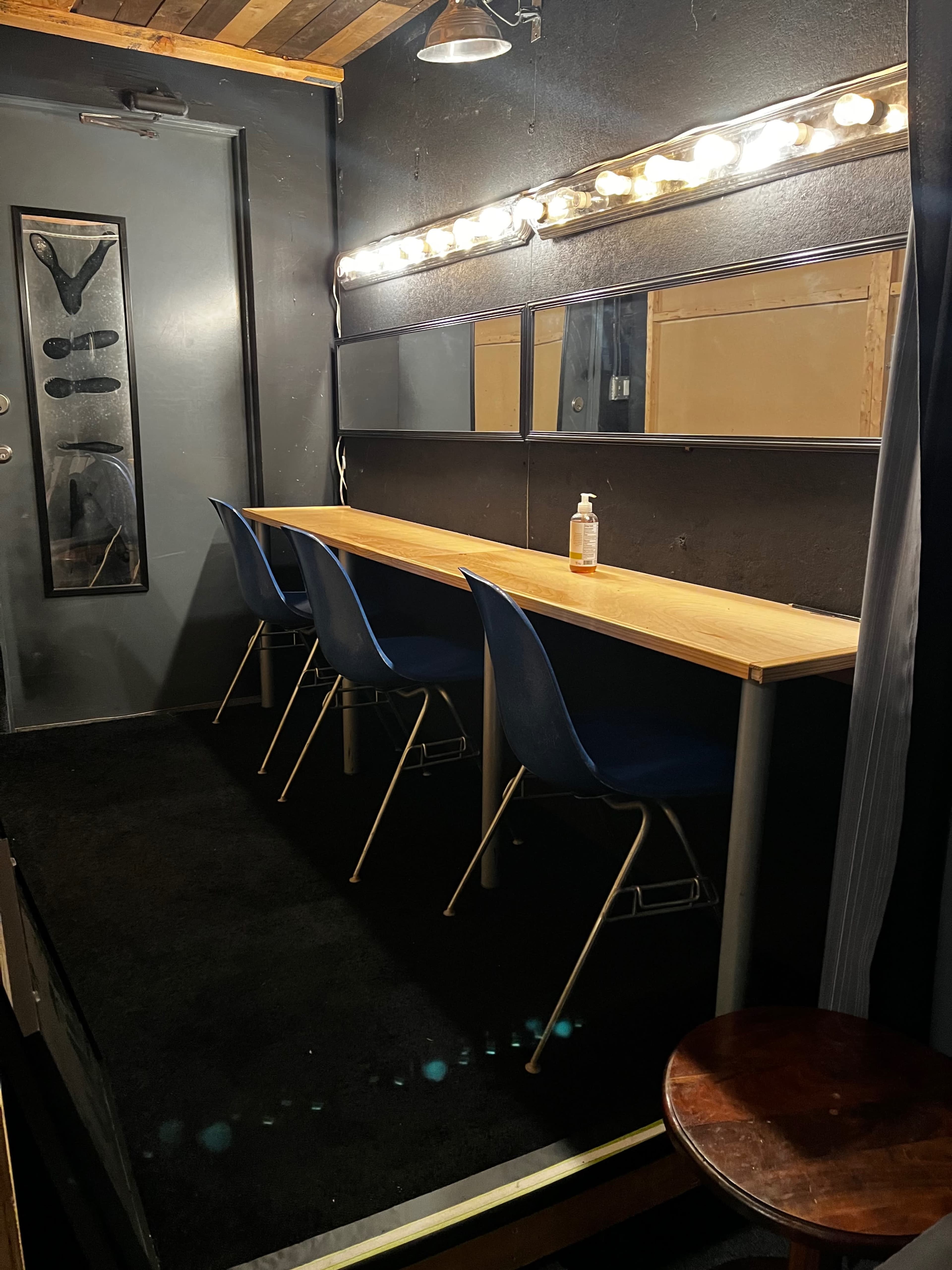 The image shows a dimly lit dressing room with a long wooden table, three blue chairs, and a mirror illuminated by light bulbs.