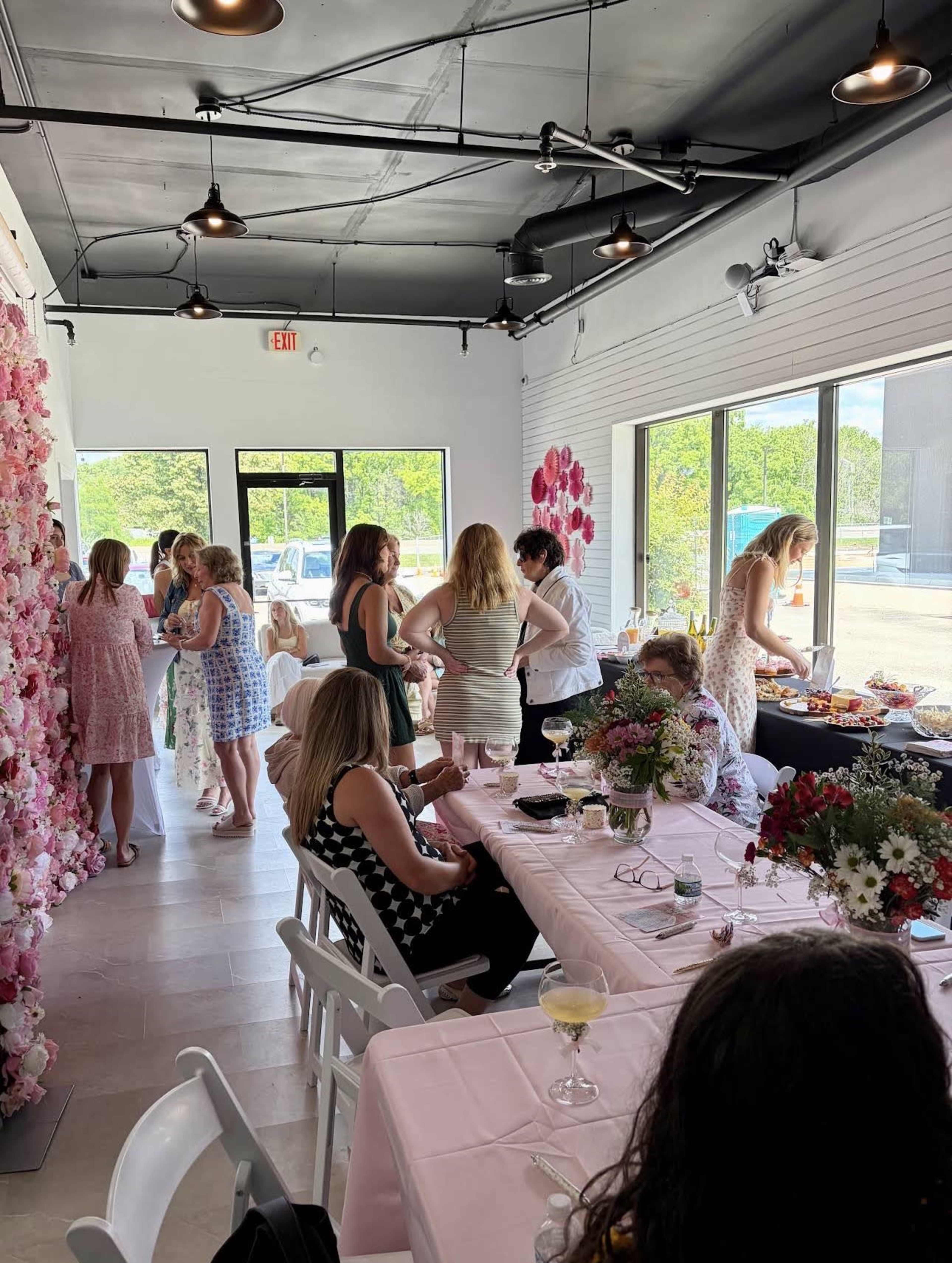 A group of people is gathered in a bright room decorated with floral arrangements, engaged in conversations and enjoying food and drinks at tables covered with pink tablecloths.