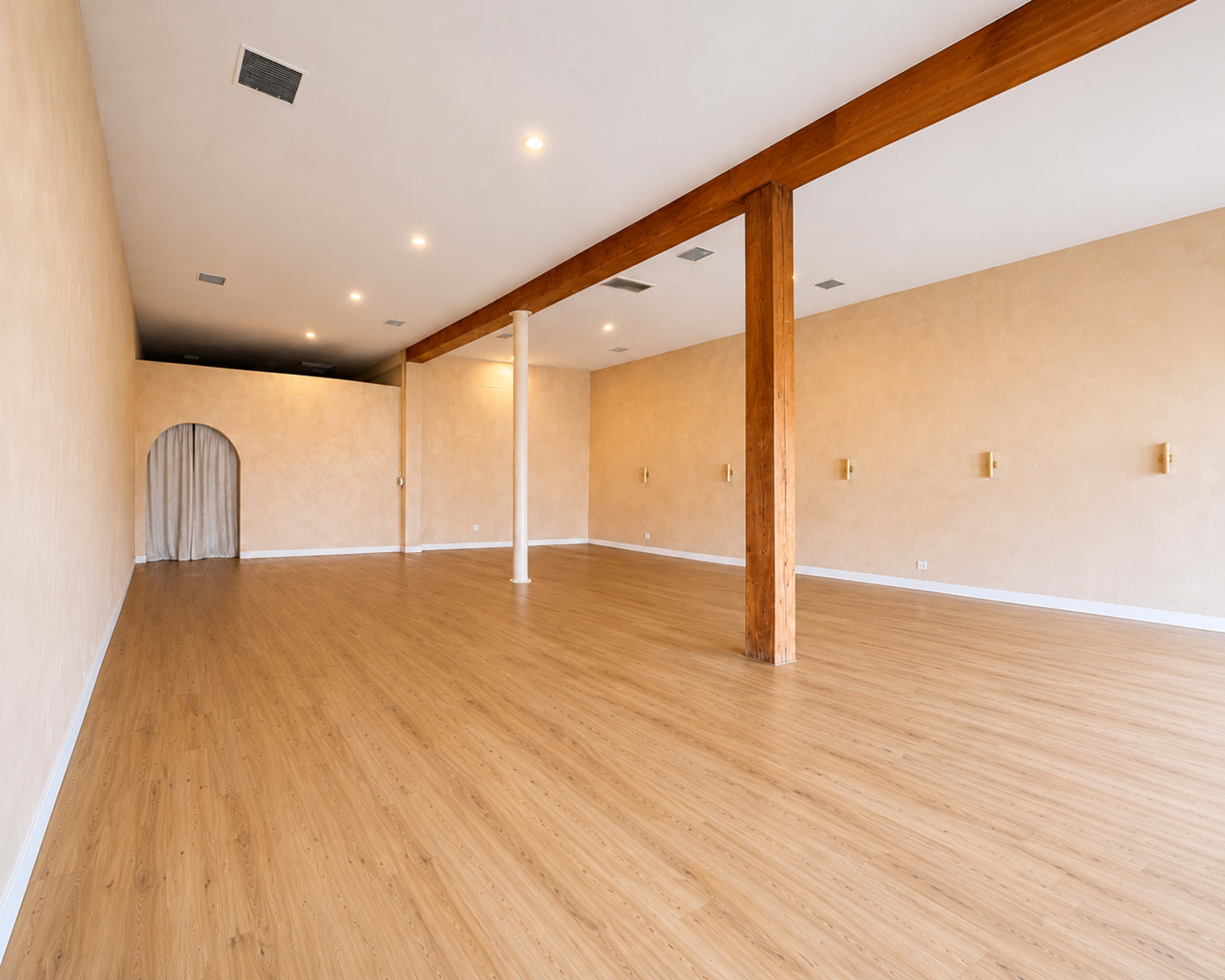 The image depicts a spacious, empty room with wooden flooring, featuring a textured beige wall and a single decorative archway.