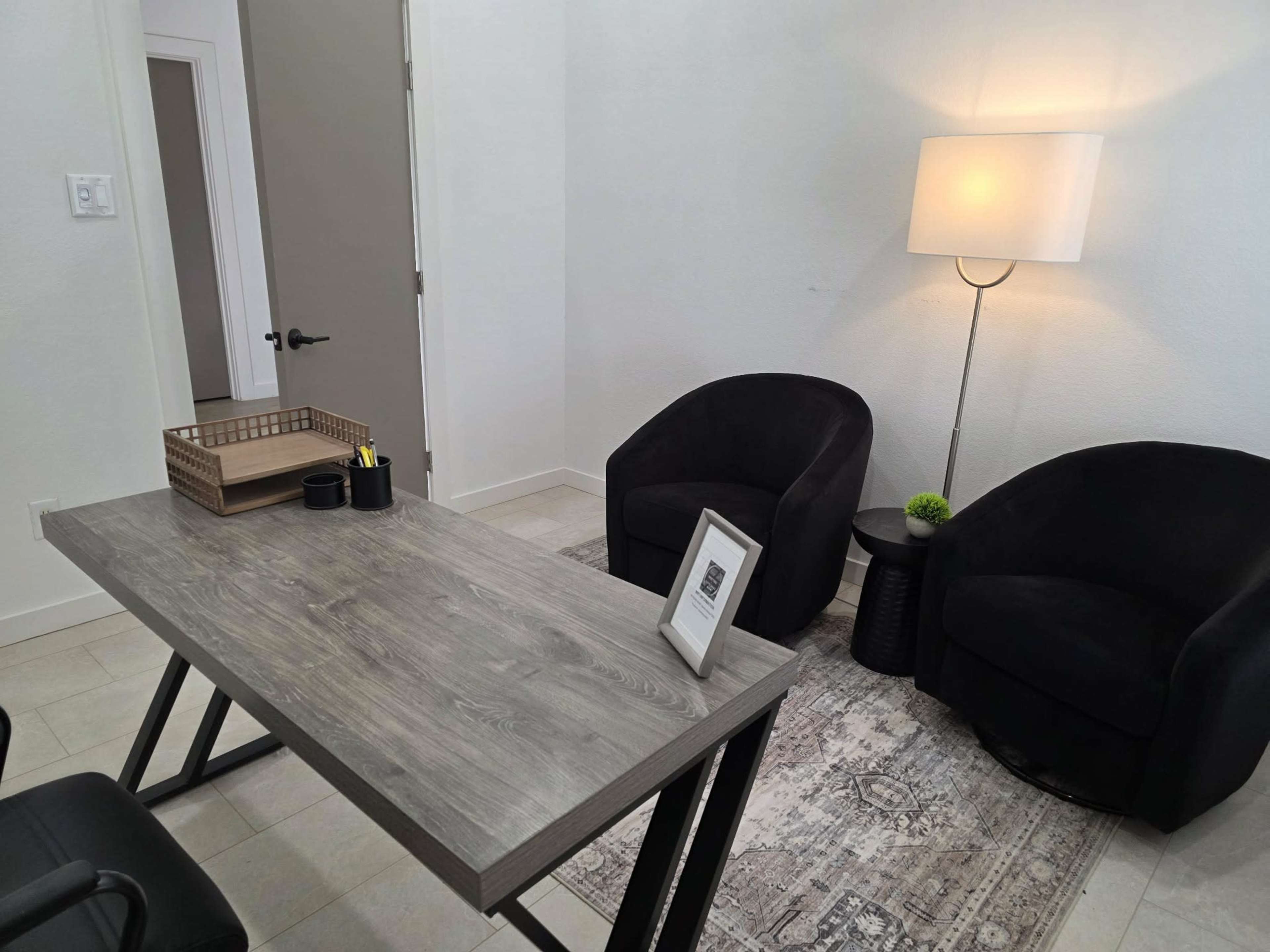 The image shows a modern office space featuring a wooden desk, a lamp, two black chairs, and a decorative rug.