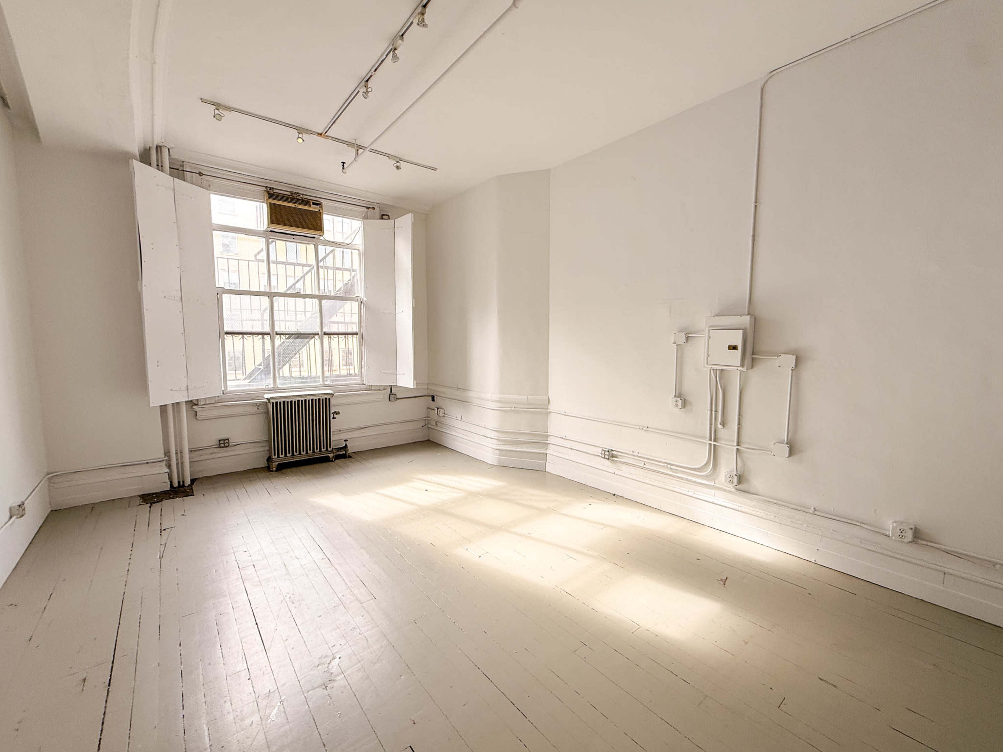 The image shows an empty room with white walls, a wooden floor, and large windows allowing natural light to enter.