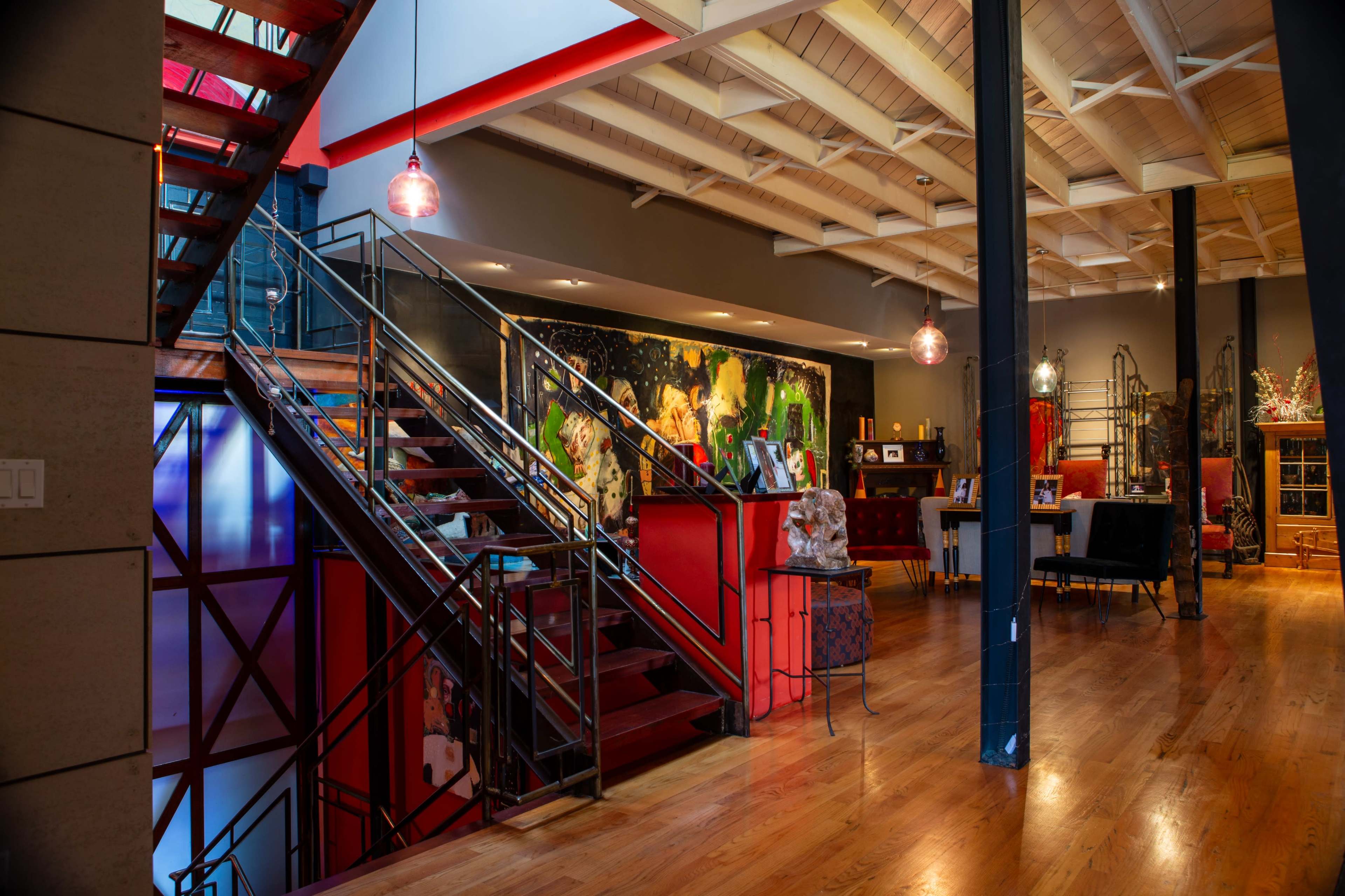 The image shows a modern interior space with a staircase leading to a second level, featuring colorful artwork on the walls and various seating areas.