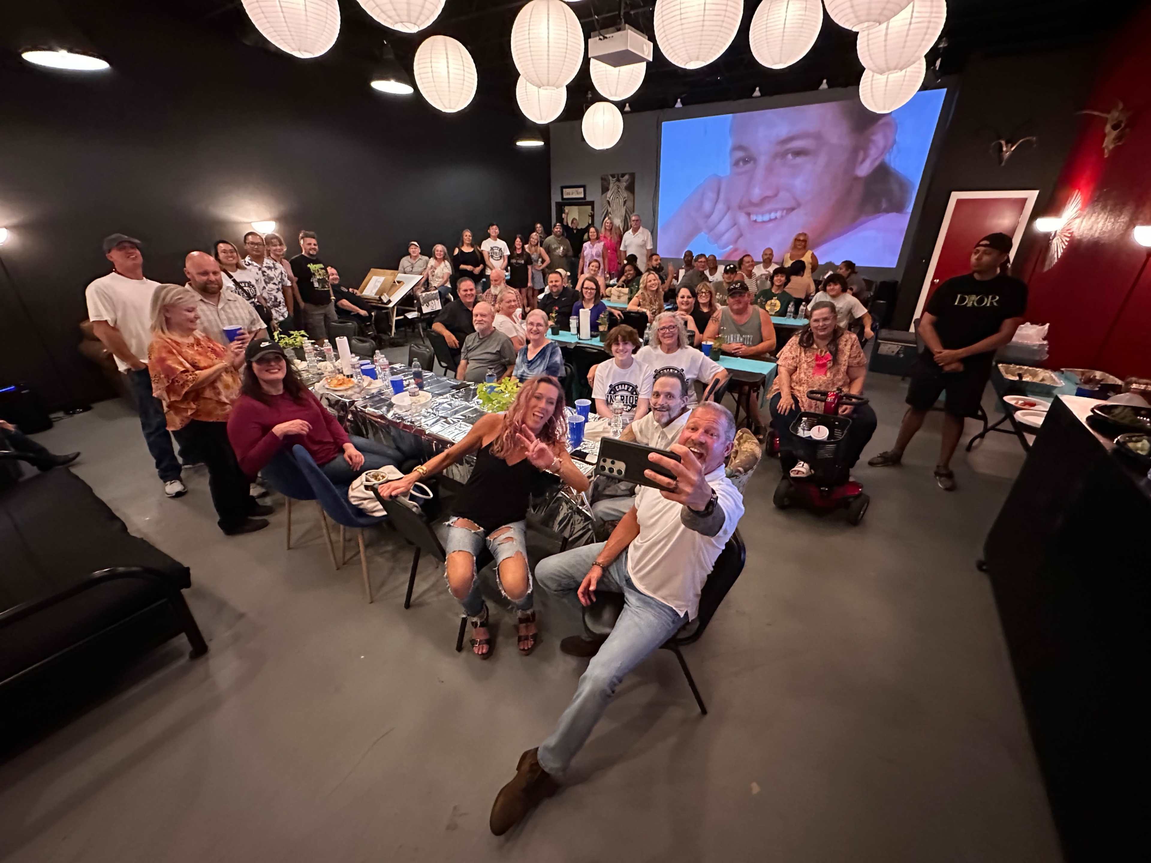 A large group of people gathers for a celebration in a dimly lit room, with a projector screen displaying a smiling face in the background.