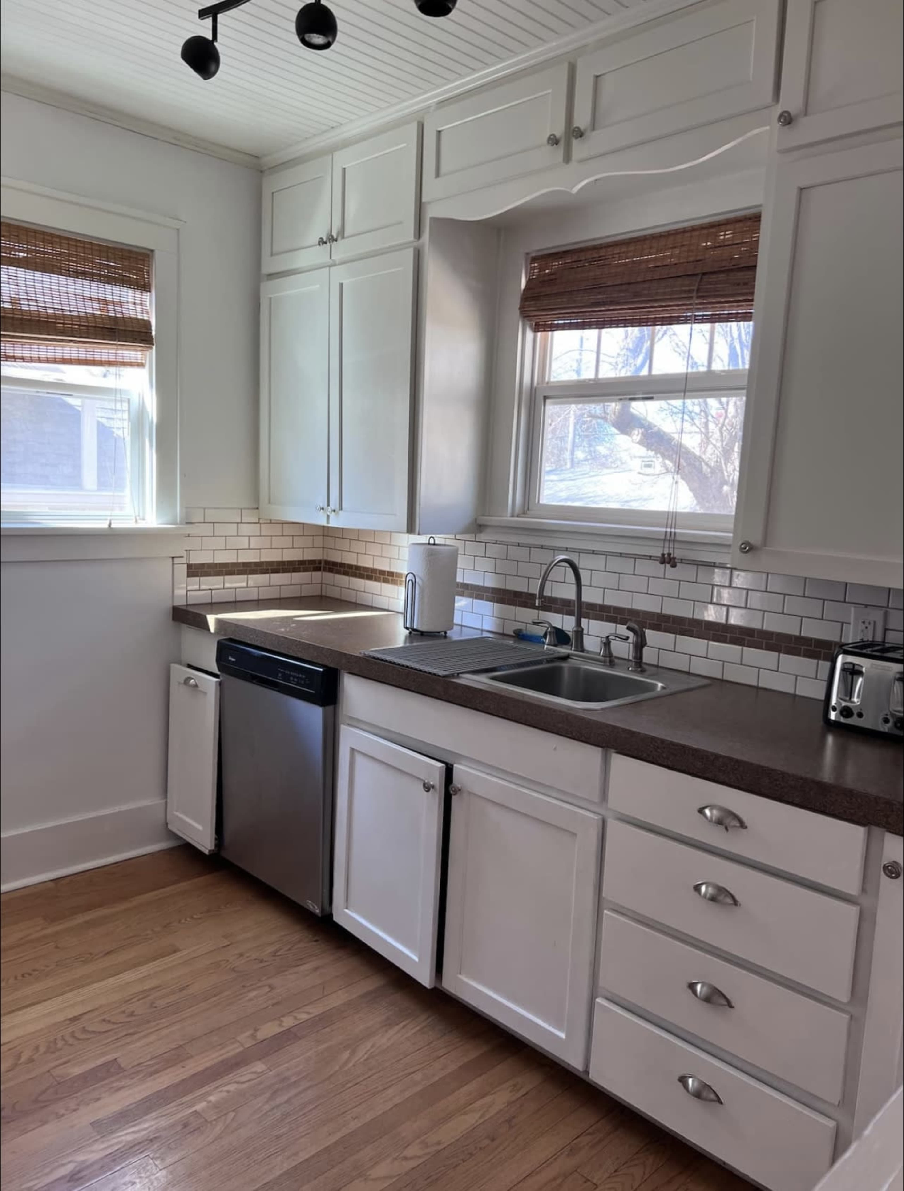 A kitchen with white cabinetry, a stainless steel dishwasher, a double sink, and a countertop with light brown accents.