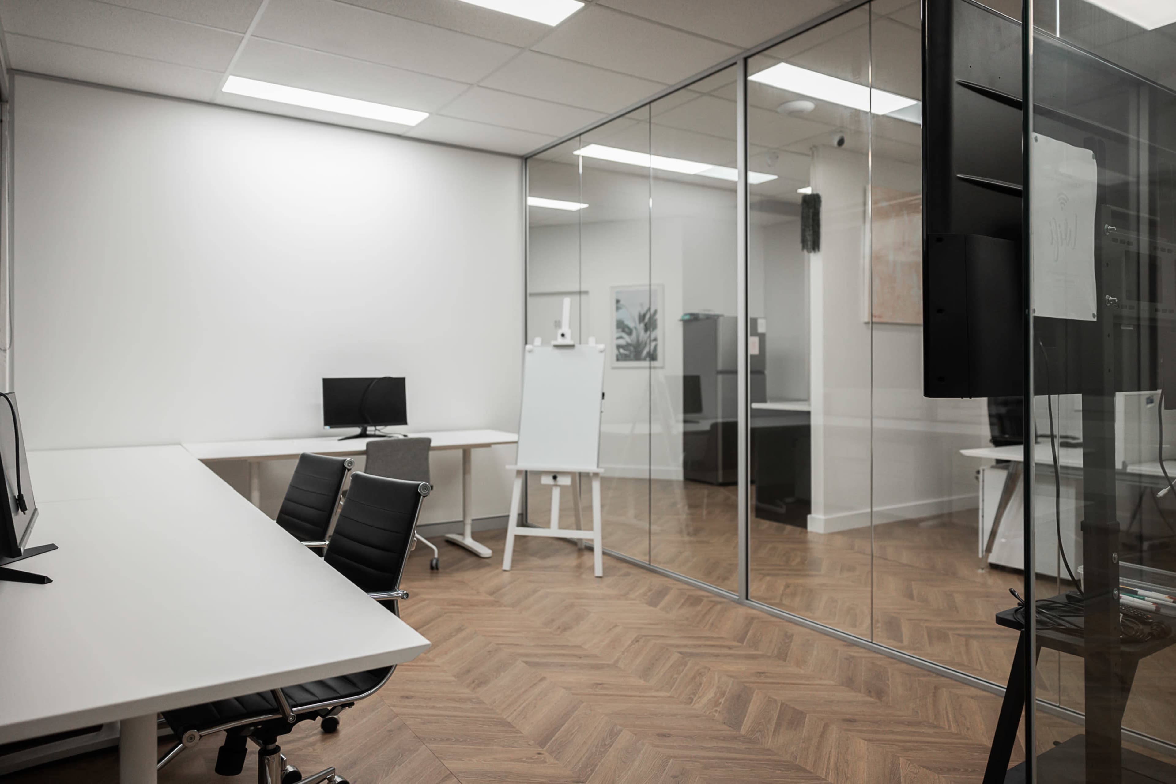 A modern office space with a glass-walled meeting room, two workstations with computers, and a whiteboard.