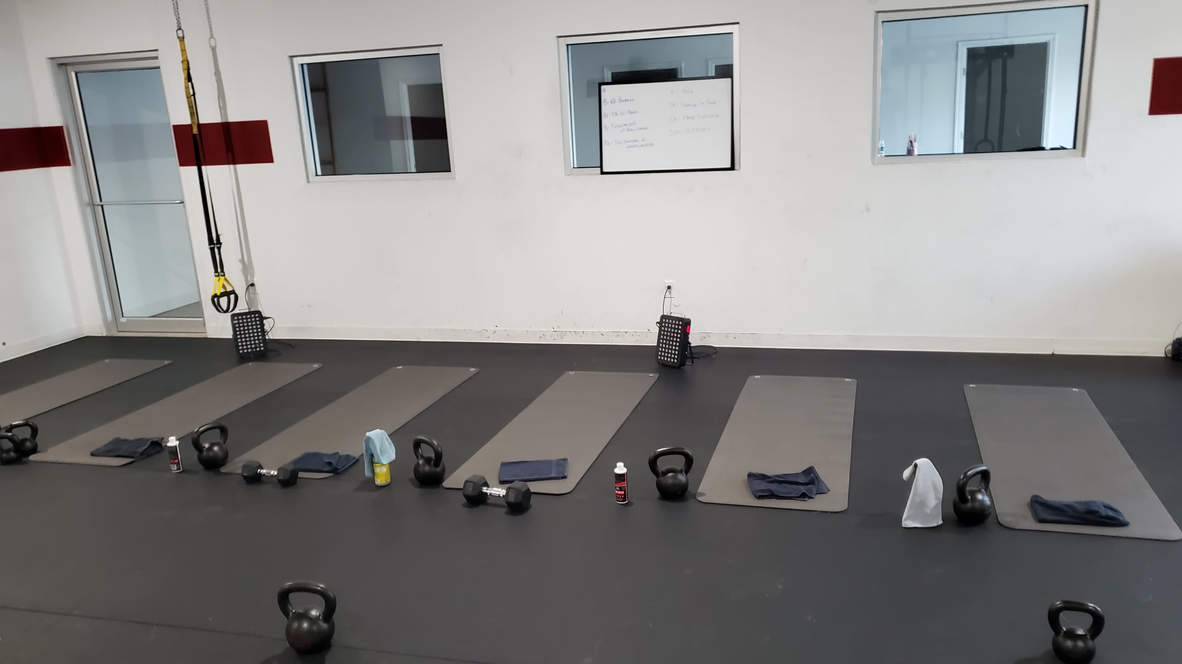 A fitness studio with several workout mats arranged in a line, each equipped with kettlebells and towels, and a whiteboard visible in the background.