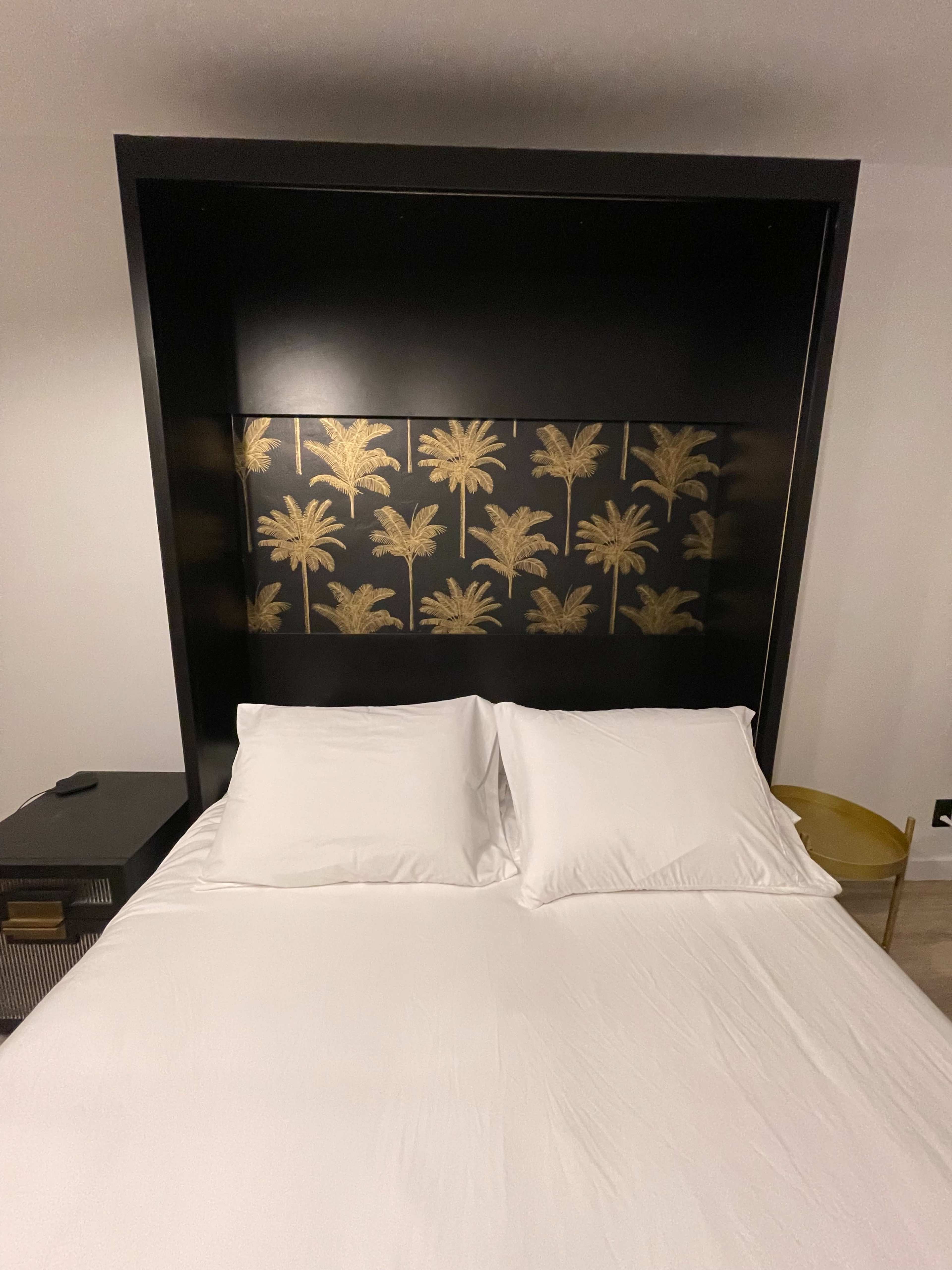 The image shows a bed with white linens and a black headboard featuring a gold palm tree design.