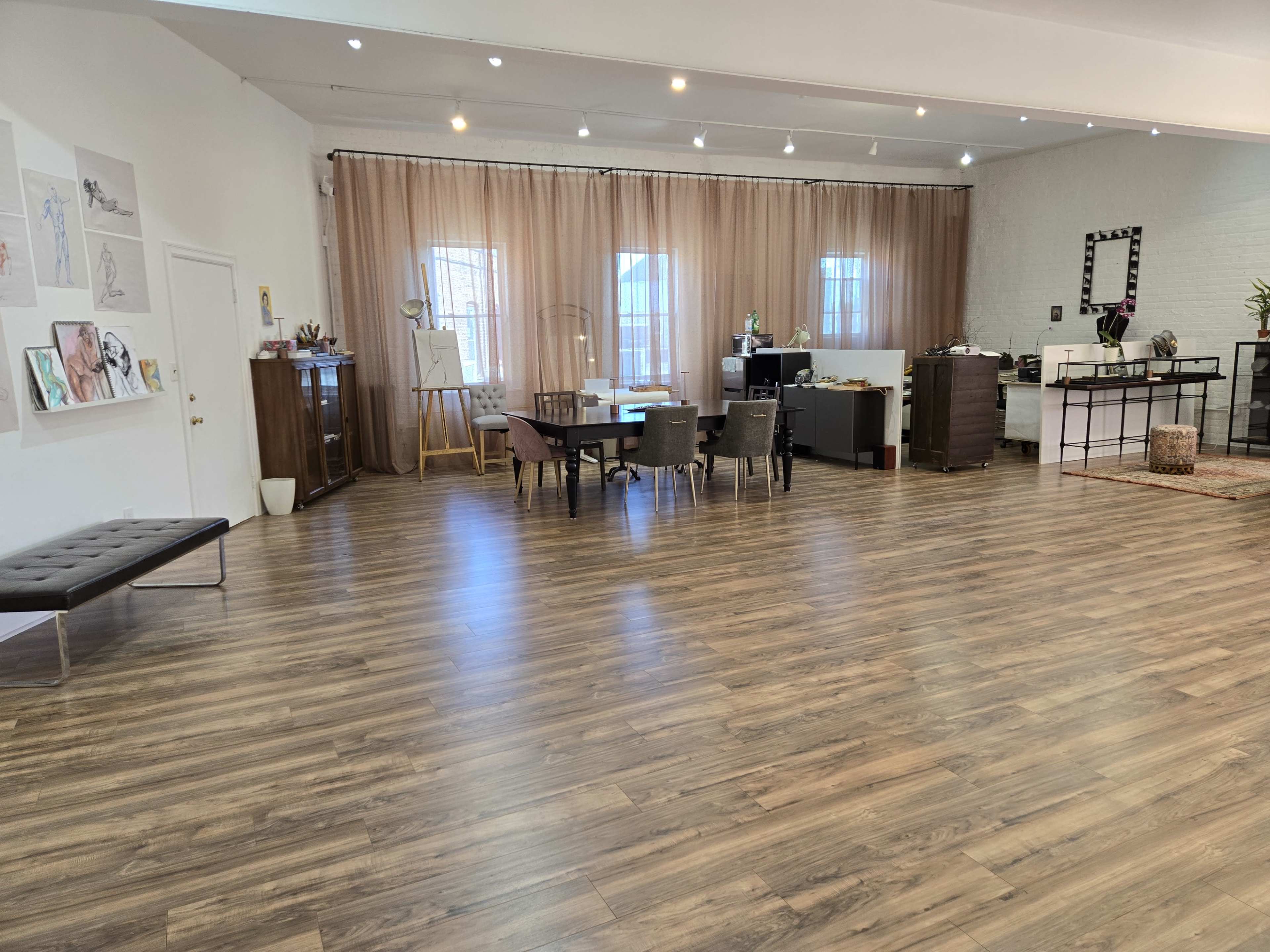 A spacious interior room with wooden flooring, featuring a mix of chairs and tables, large windows draped with curtains, and various artworks displayed on the walls.