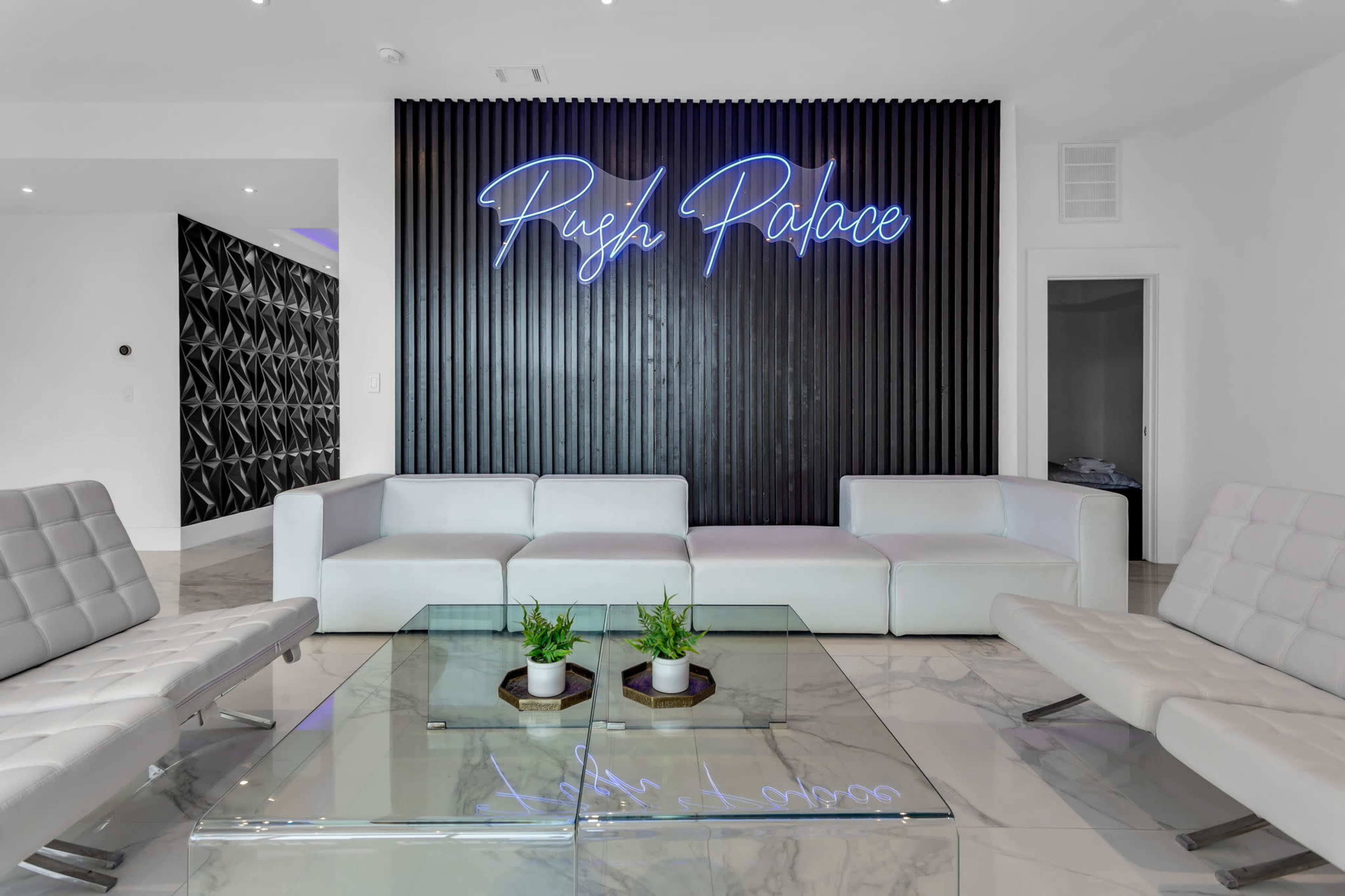 A modern waiting area with white sofas, a glass coffee table, and a neon sign that reads "Pish Palace" against a black textured wall.