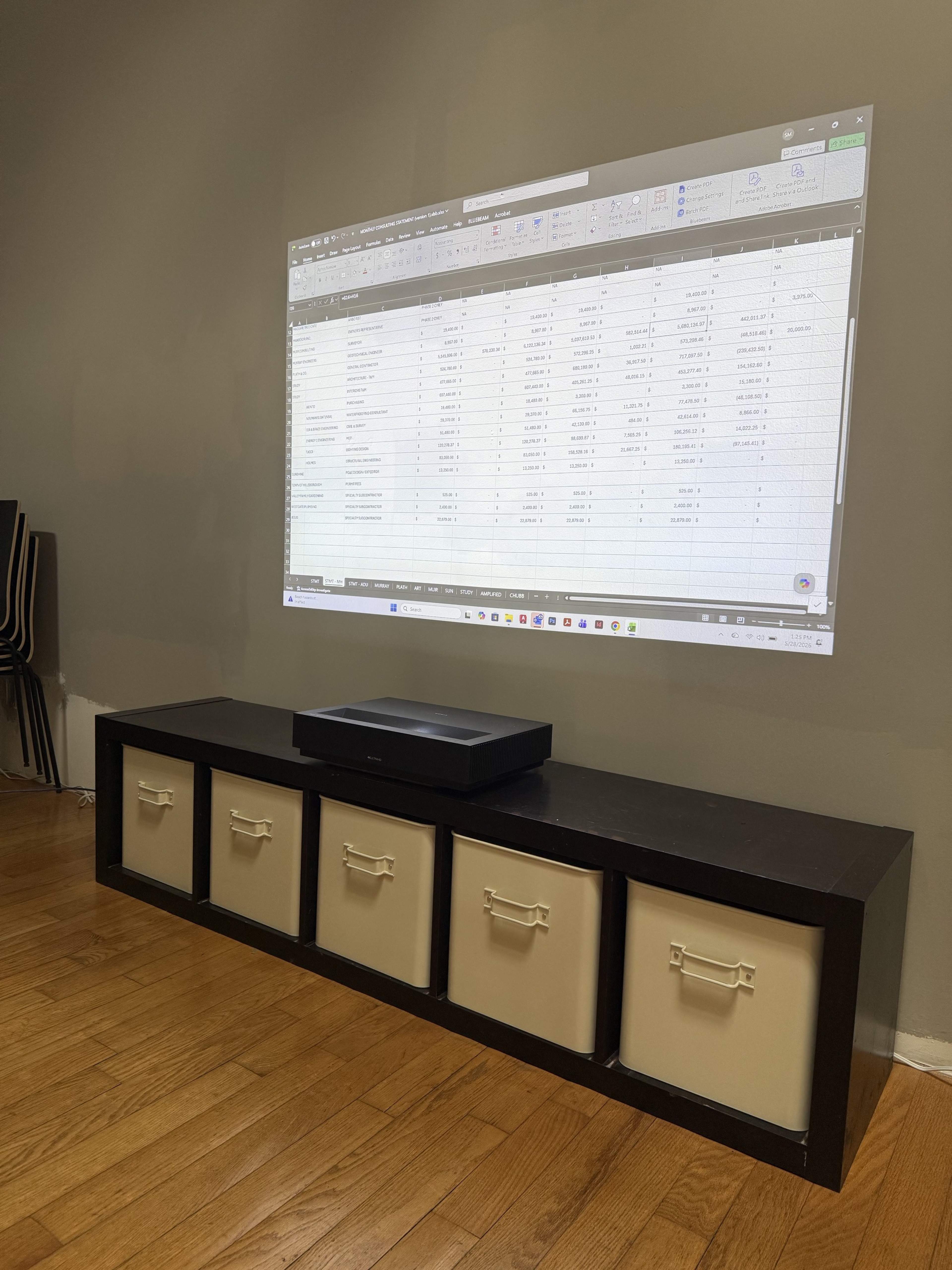 A projector casts an Excel spreadsheet onto a gray wall above a dark storage unit with four beige bins.