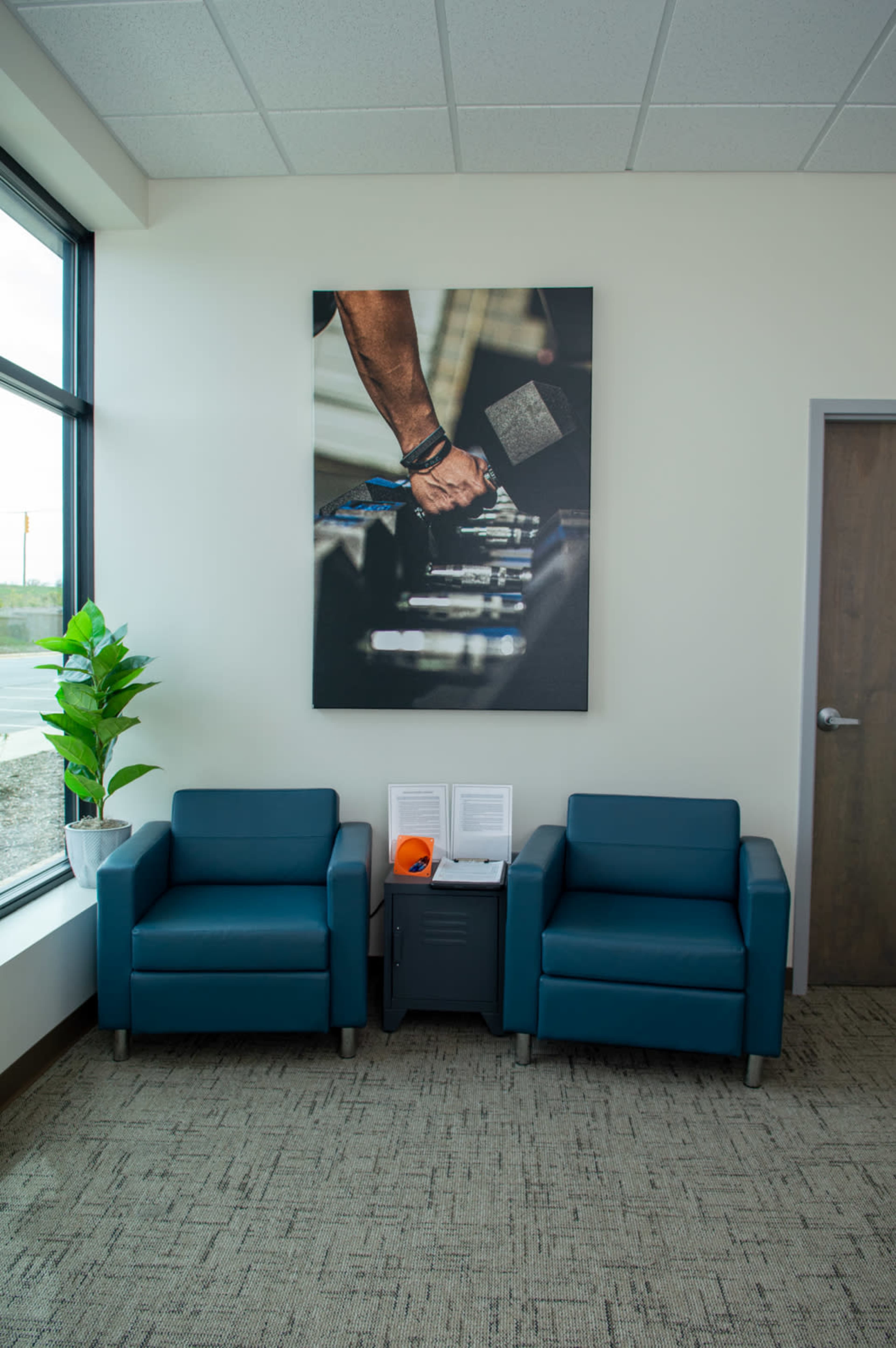 The image shows a modern waiting area with two teal chairs, a small table, a potted plant, and a large wall art piece featuring a close-up of a person's hand holding a dumbbell.