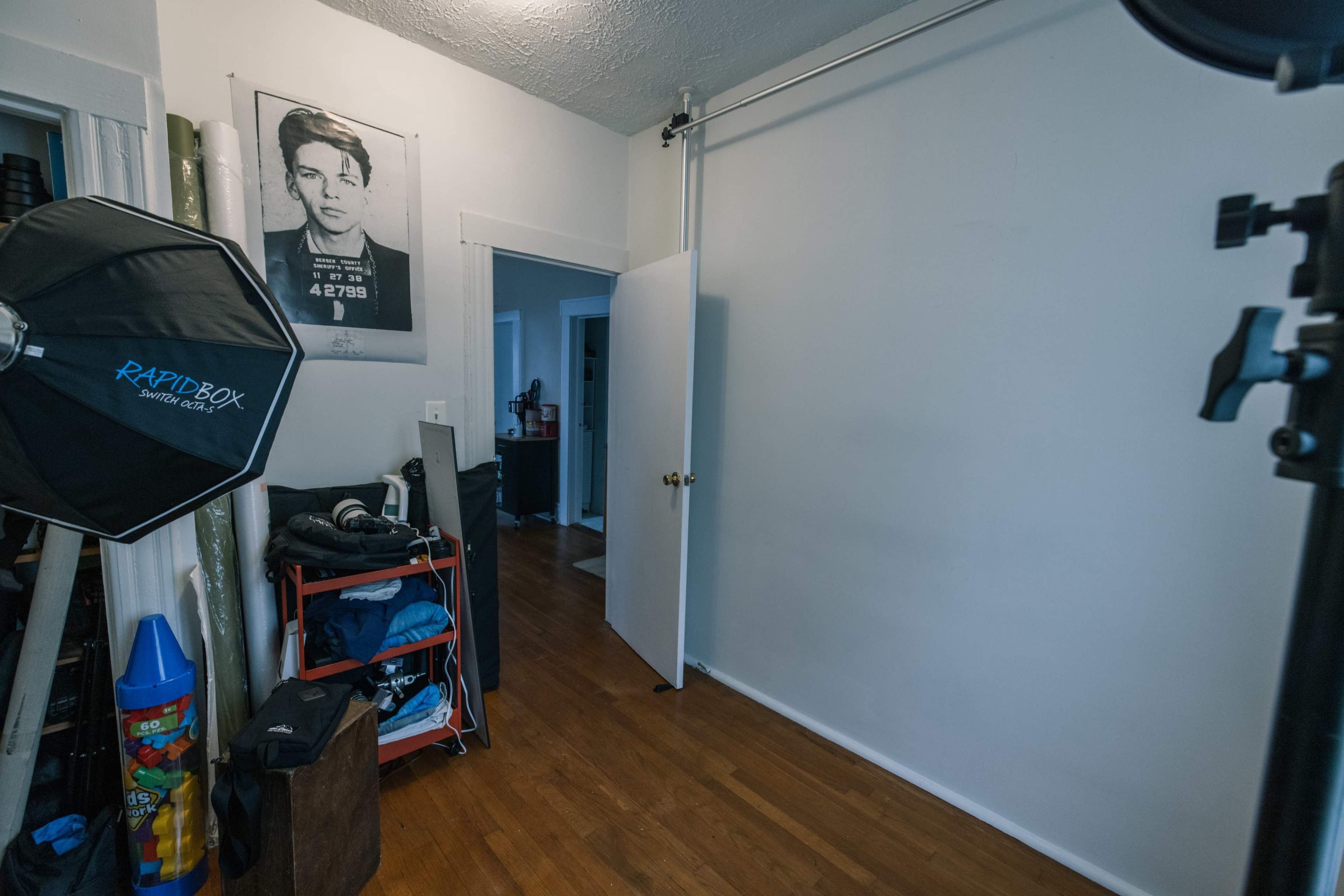 A partially furnished room with a door on the right, a photography umbrella on the left, and a wall featuring a large portrait above a shelf.