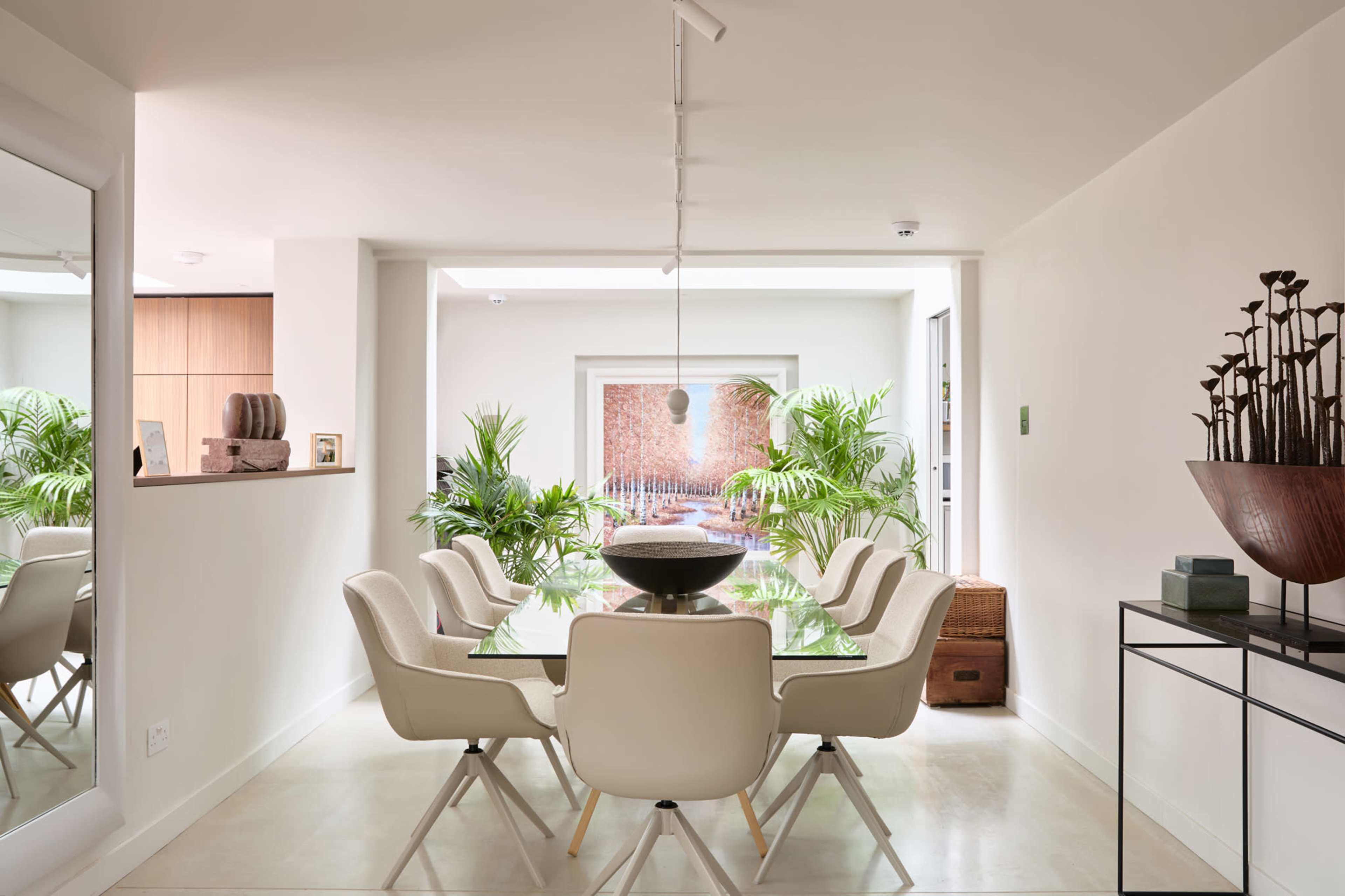 A modern dining area features a glass table surrounded by white chairs, with plants and a view of an outdoor space framed by large windows.