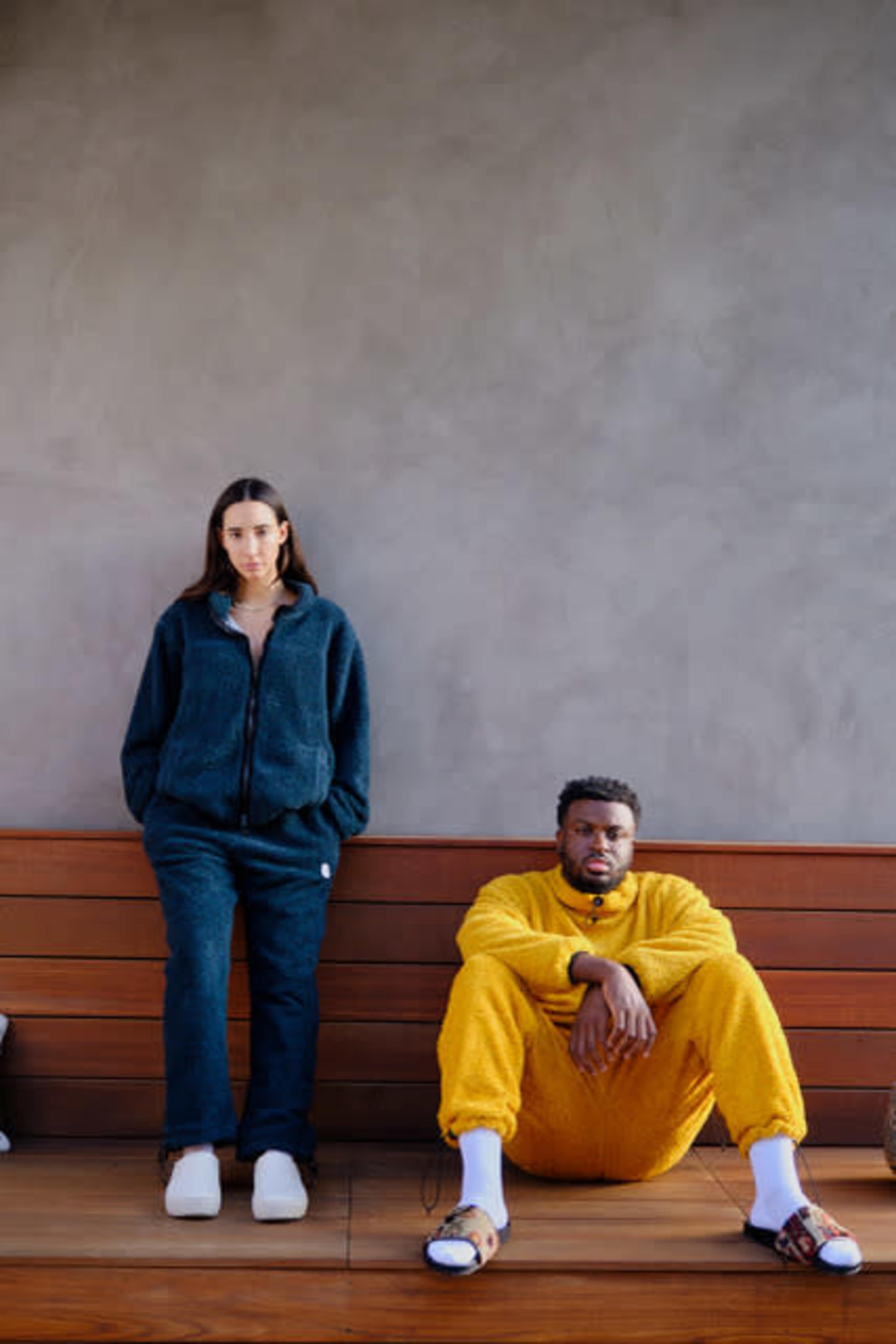 A woman in a dark outfit stands beside a man sitting on a wooden bench, who is dressed in a bright yellow onesie.