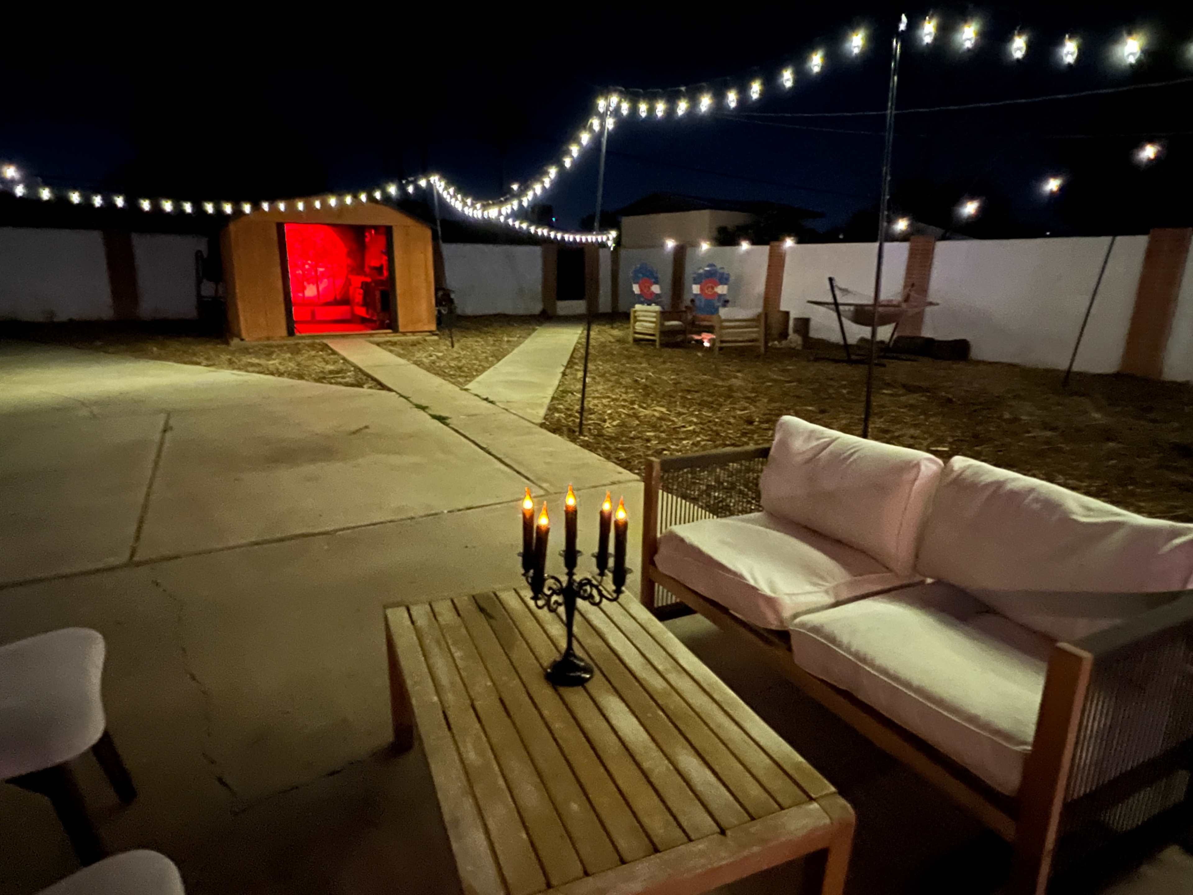 The image shows a backyard at night with string lights overhead, a wooden shed in the background, and a seating area featuring a candle holder on a wooden table and a light-colored couch.