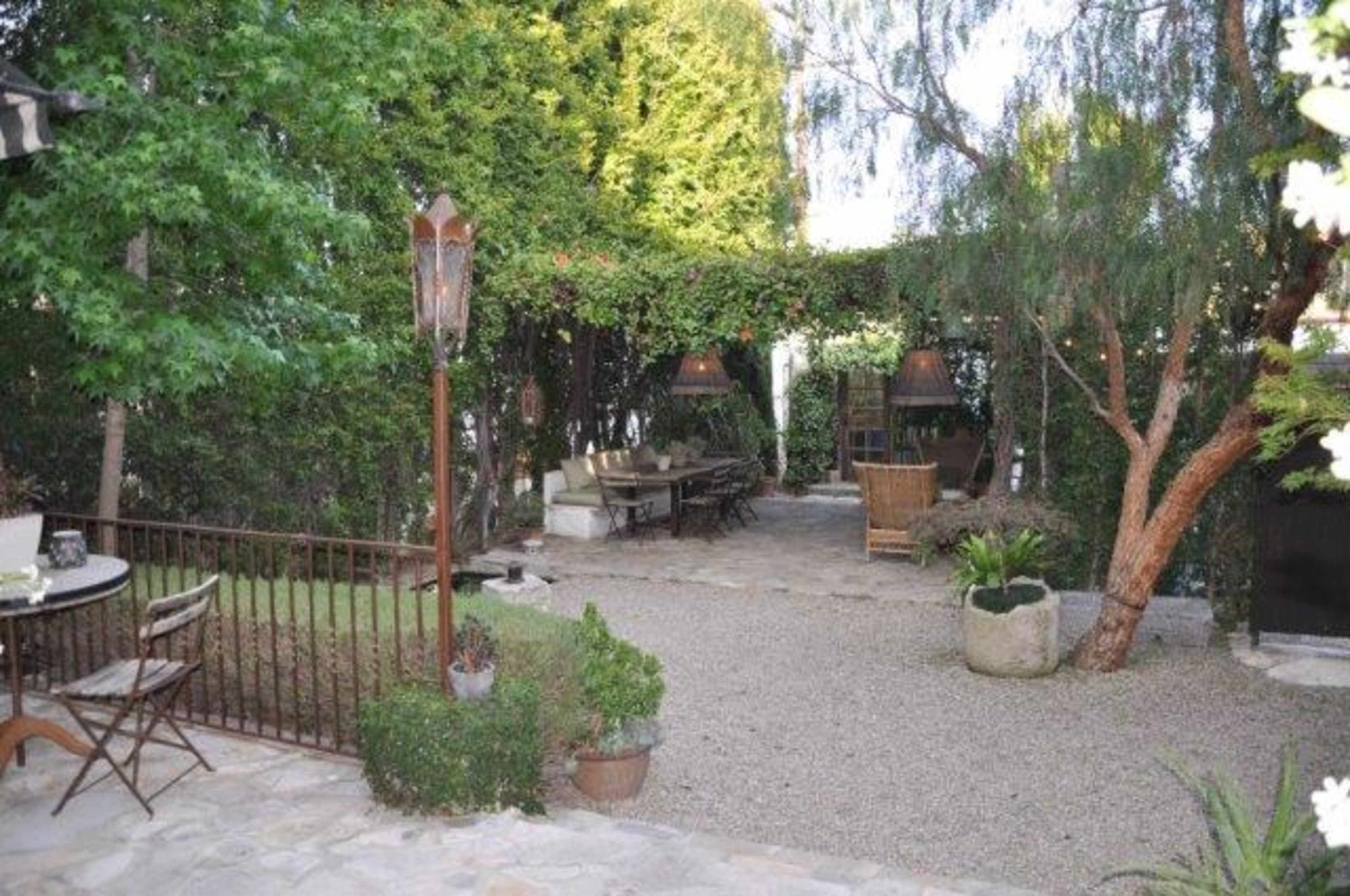 A landscaped garden features a gravel pathway, a table with chairs, and a shaded seating area surrounded by greenery.