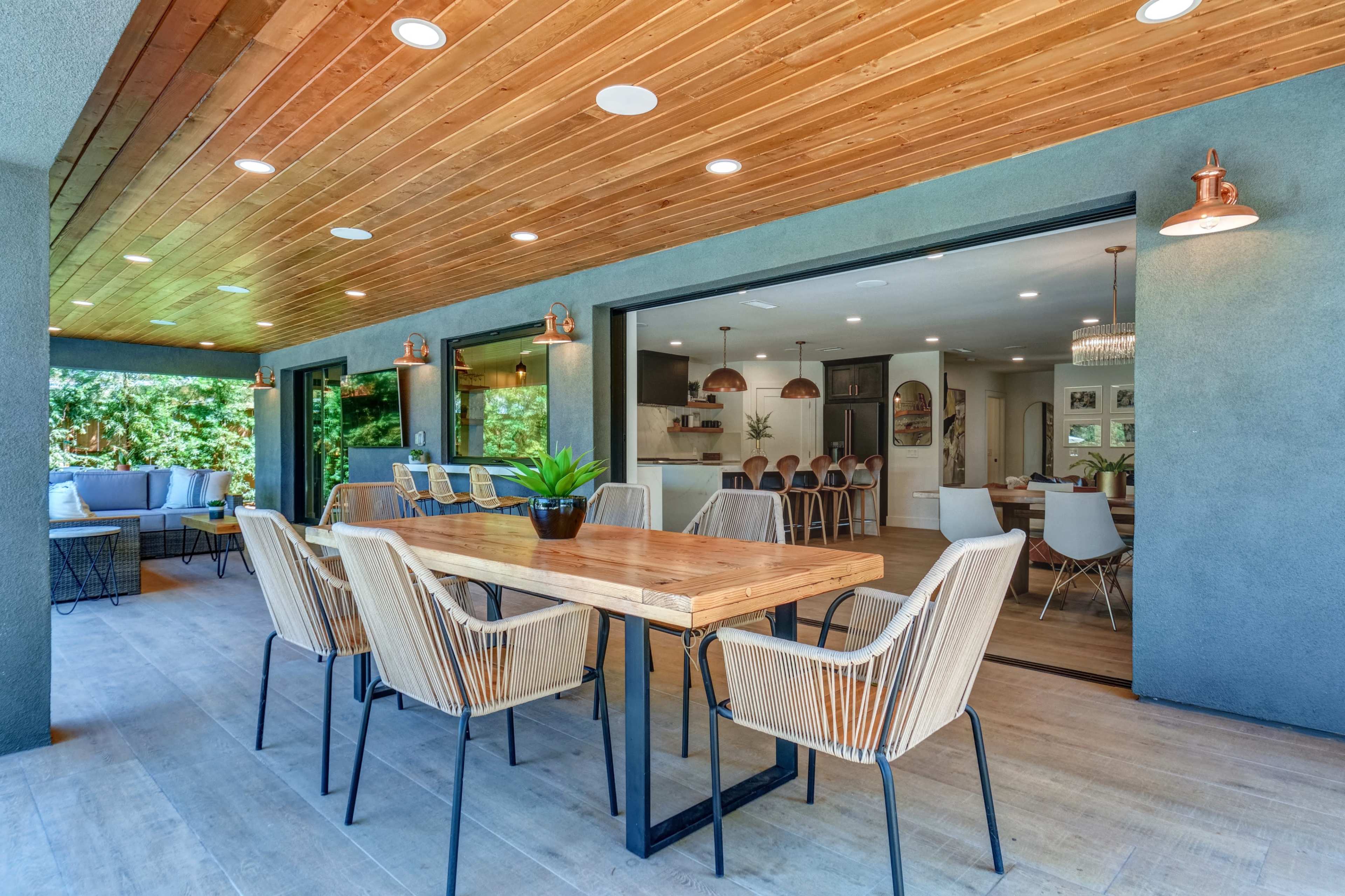 A spacious outdoor dining area features a wooden table surrounded by woven chairs, set under a wooden ceiling with recessed lighting.