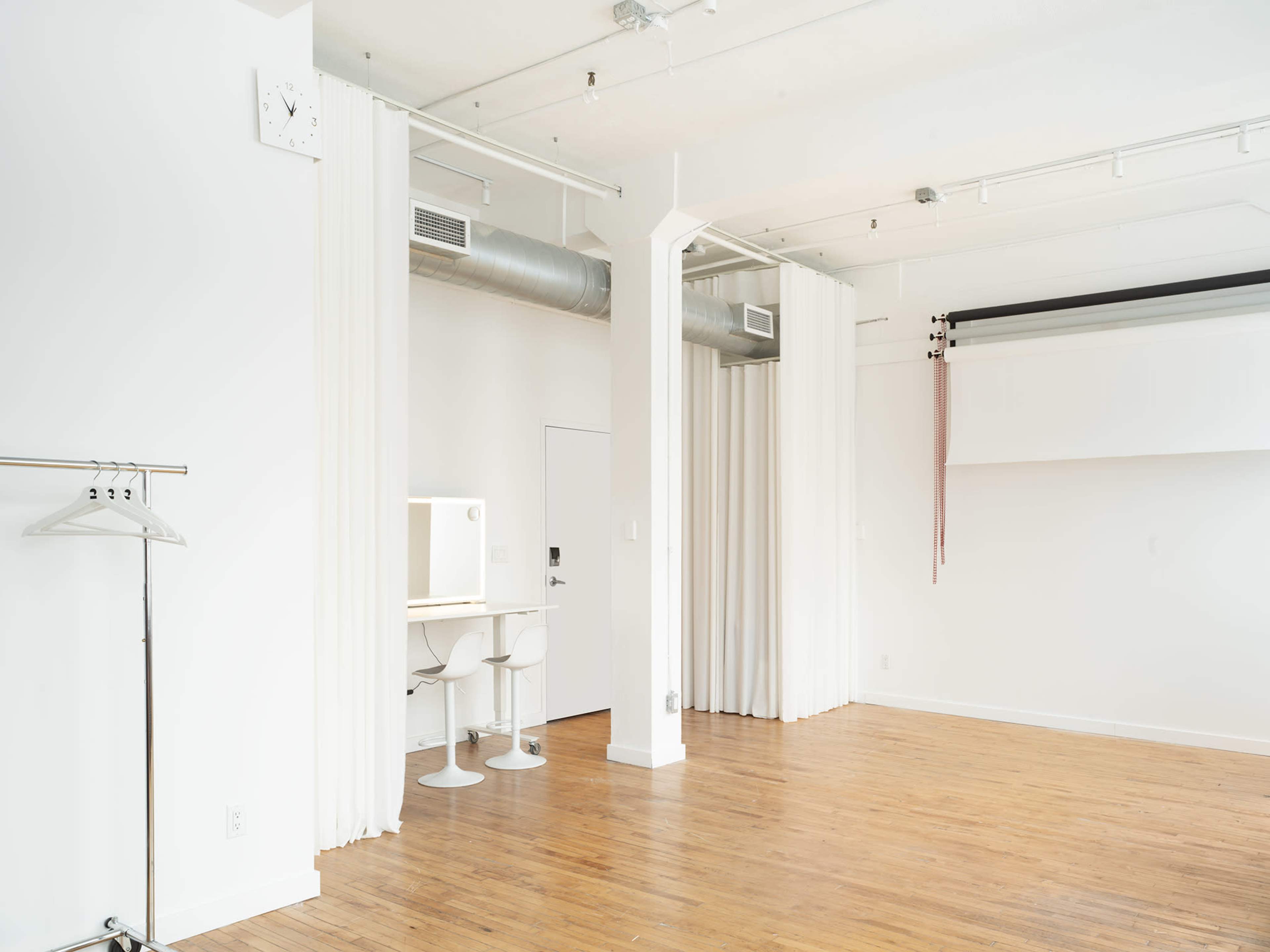 The image shows a bright, minimalistic interior space with wooden floors, white walls, and a small area featuring a desk, chair, and hanging garments.