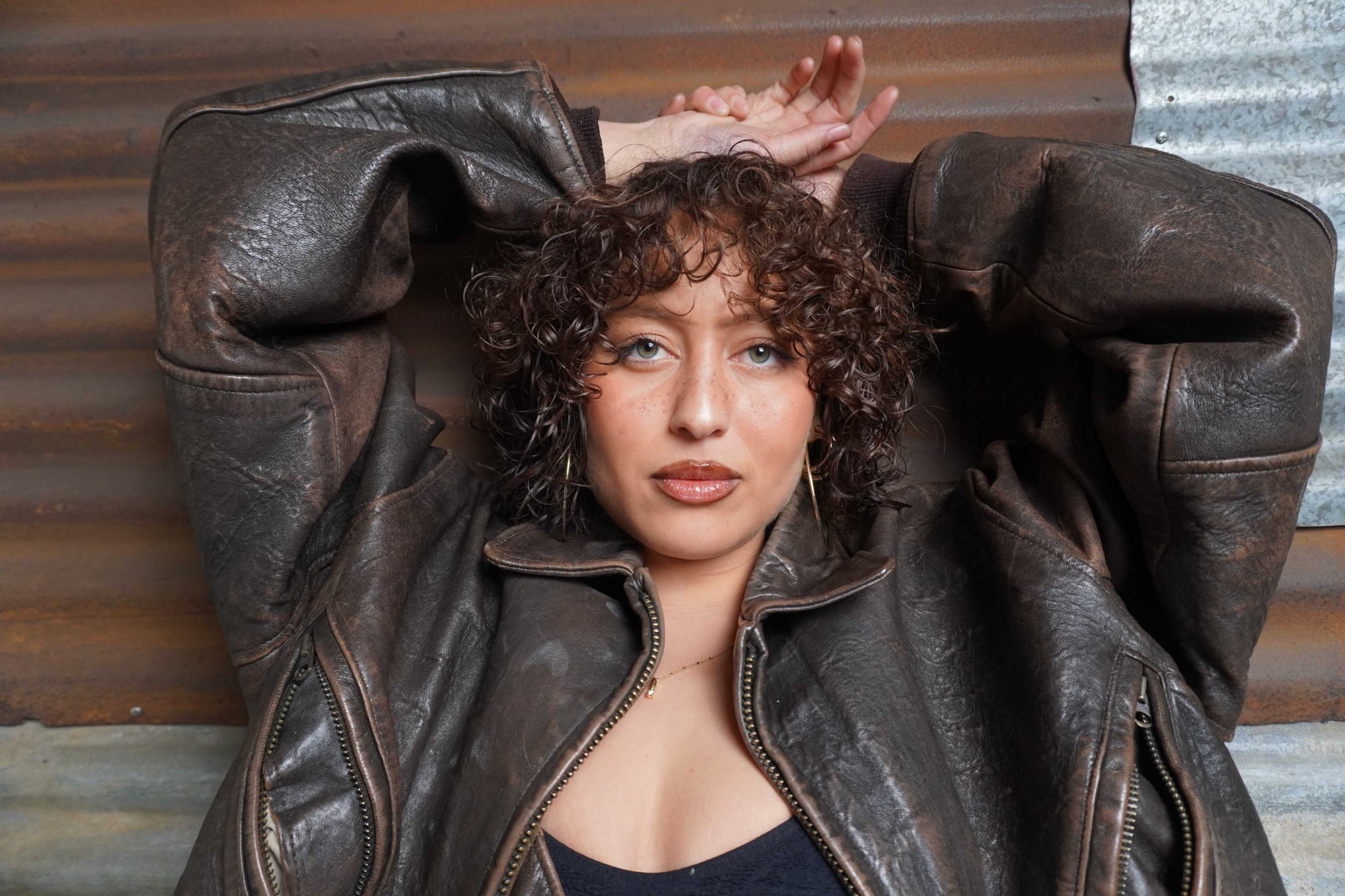 A person with curly hair poses against a textured metal wall while wearing a brown leather jacket.