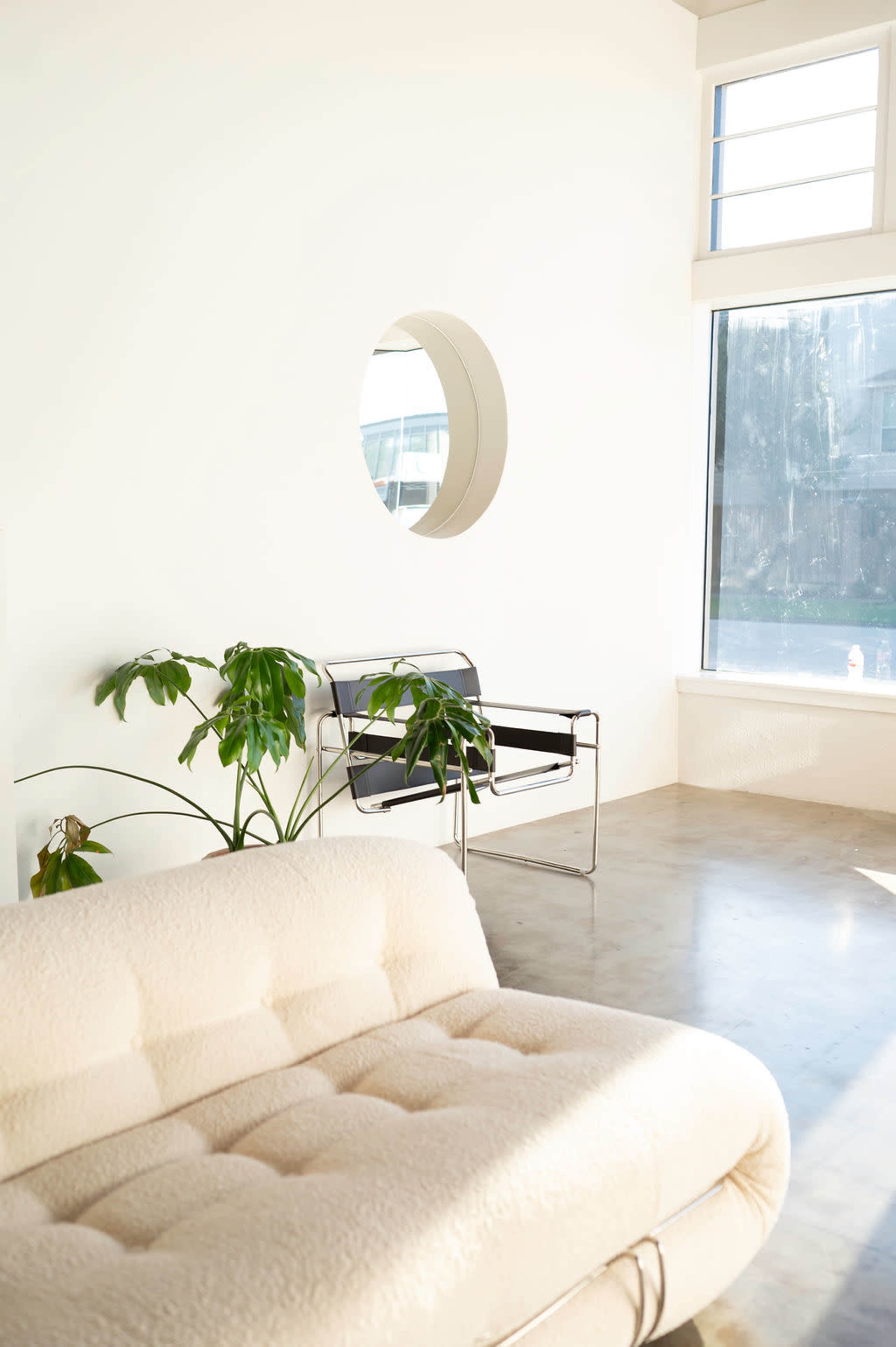 The image shows a bright, minimalist interior with a tufted white sofa, a sleek black and chrome chair, and a round mirror, complemented by a large window and a green plant.