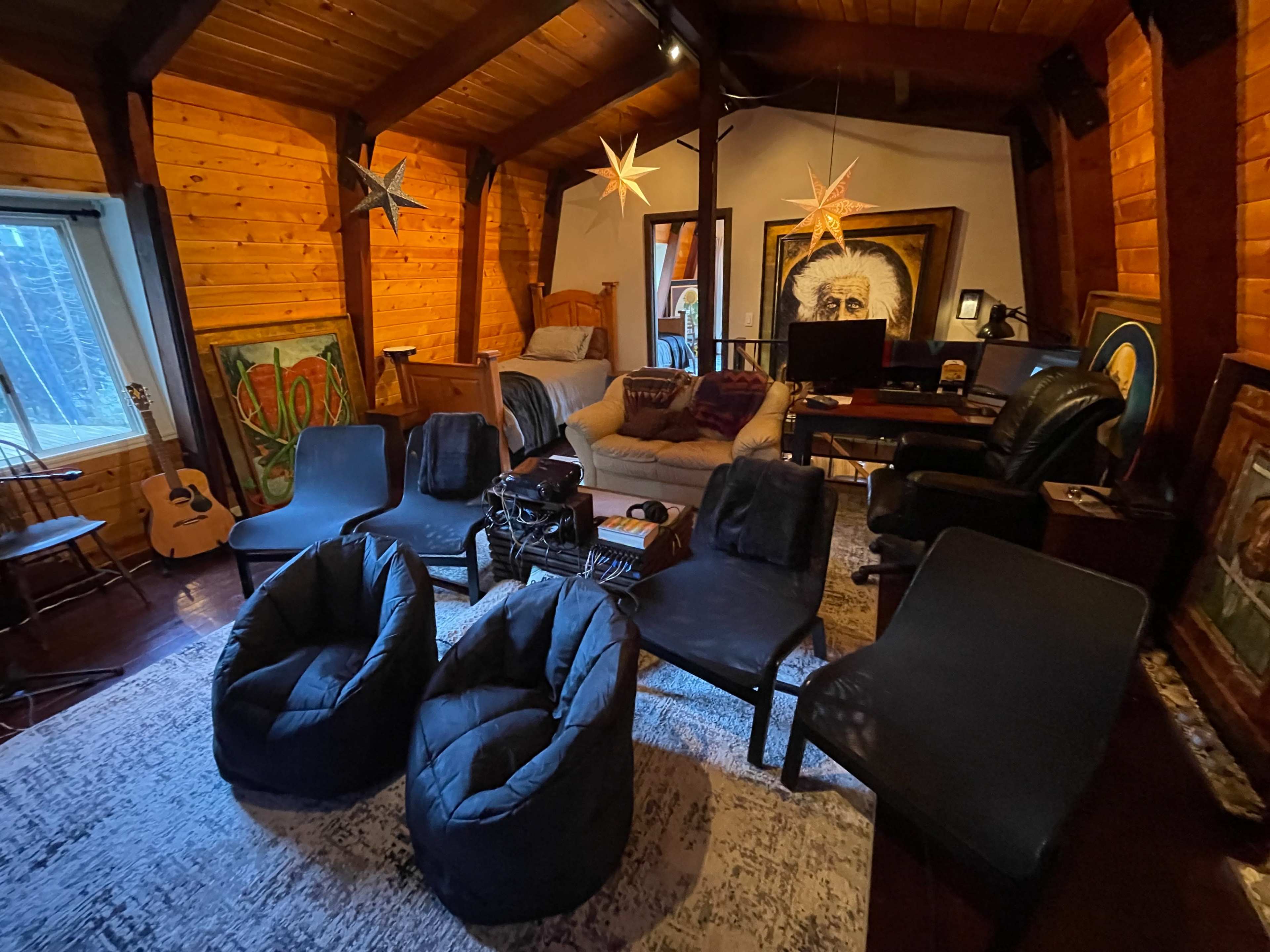 A cozy room with wooden walls featuring a seating area with bean bag chairs, a small table, artwork on the walls, and a bed in one corner.