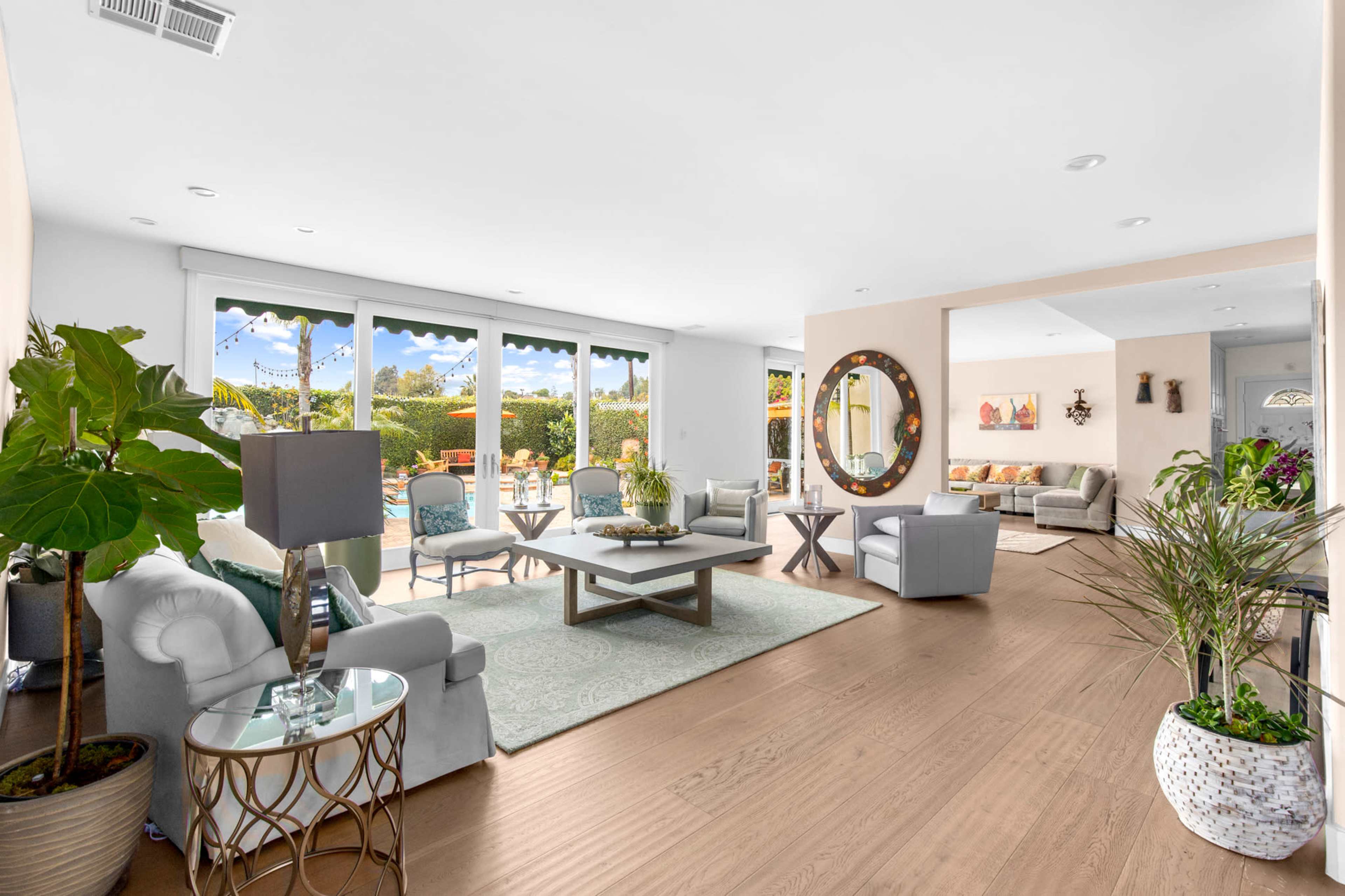 The image shows a bright, spacious living area featuring comfortable seating, a central coffee table, and large windows overlooking a landscaped outdoor space with a pool.