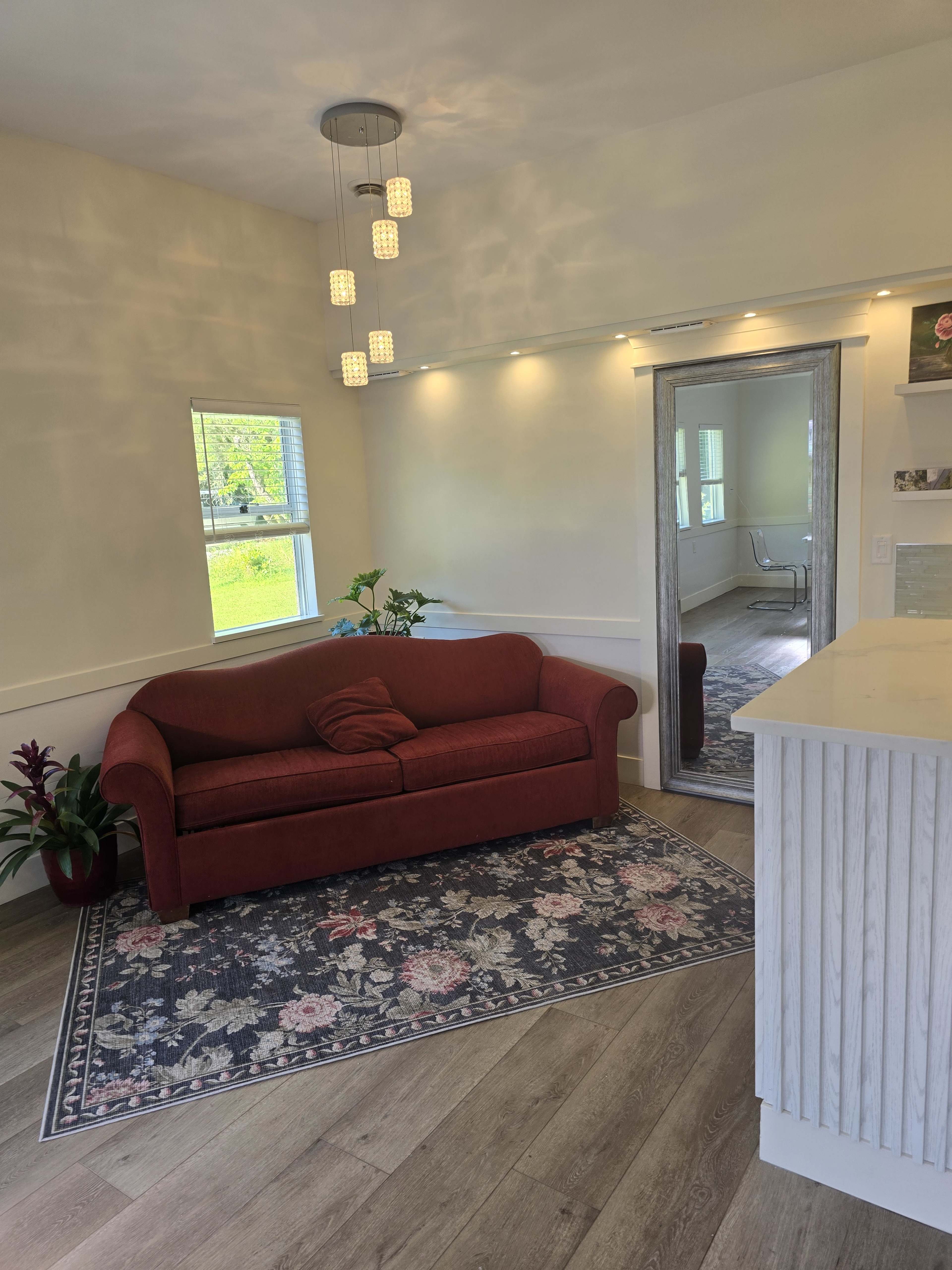 The image shows a cozy room with a red sofa, a decorative rug, a large mirror, and natural light coming through a window.