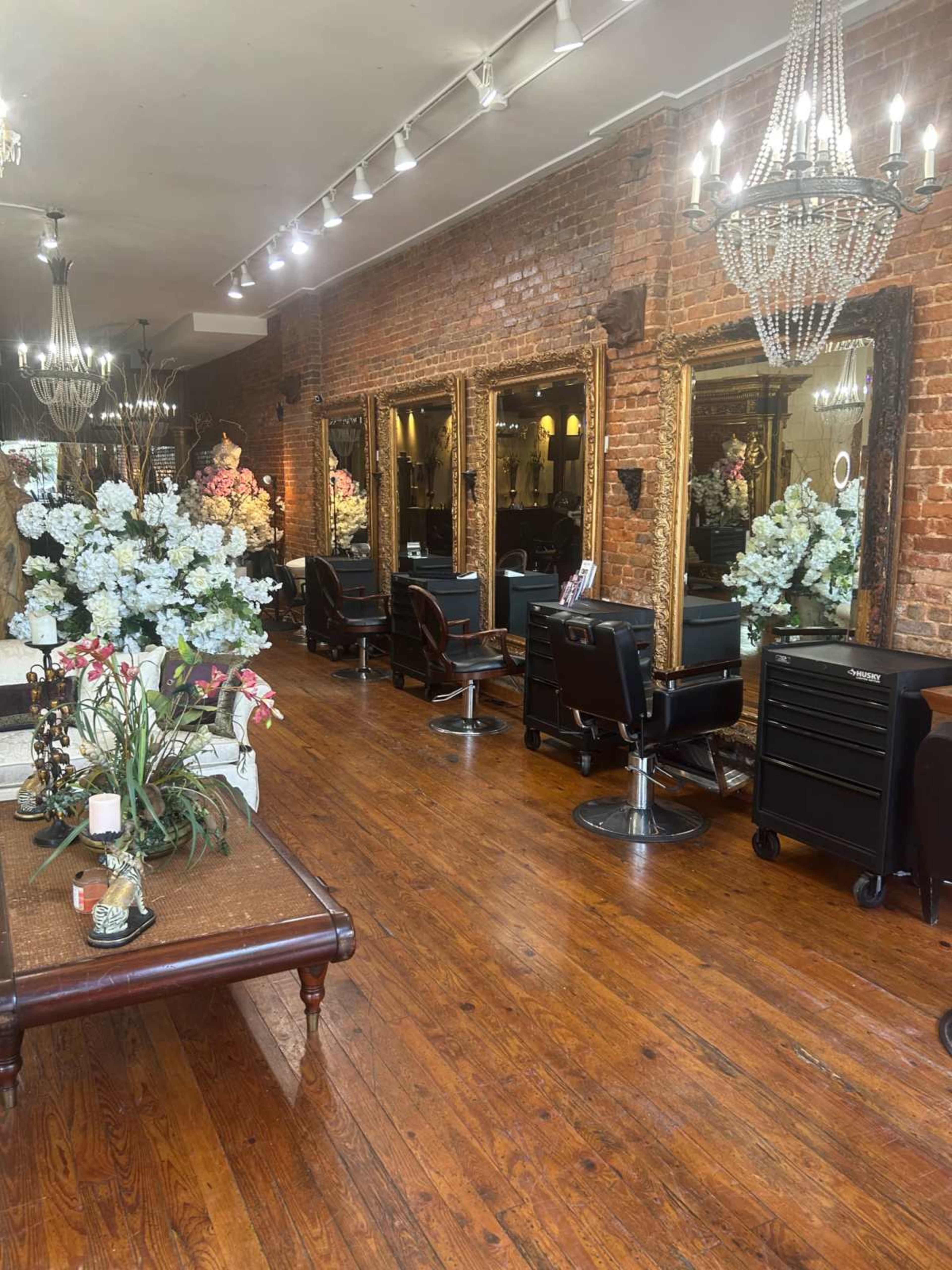 The image shows a salon interior with hardwood floors, large mirrors, and several styling chairs arranged across the space, decorated with artificial flowers.