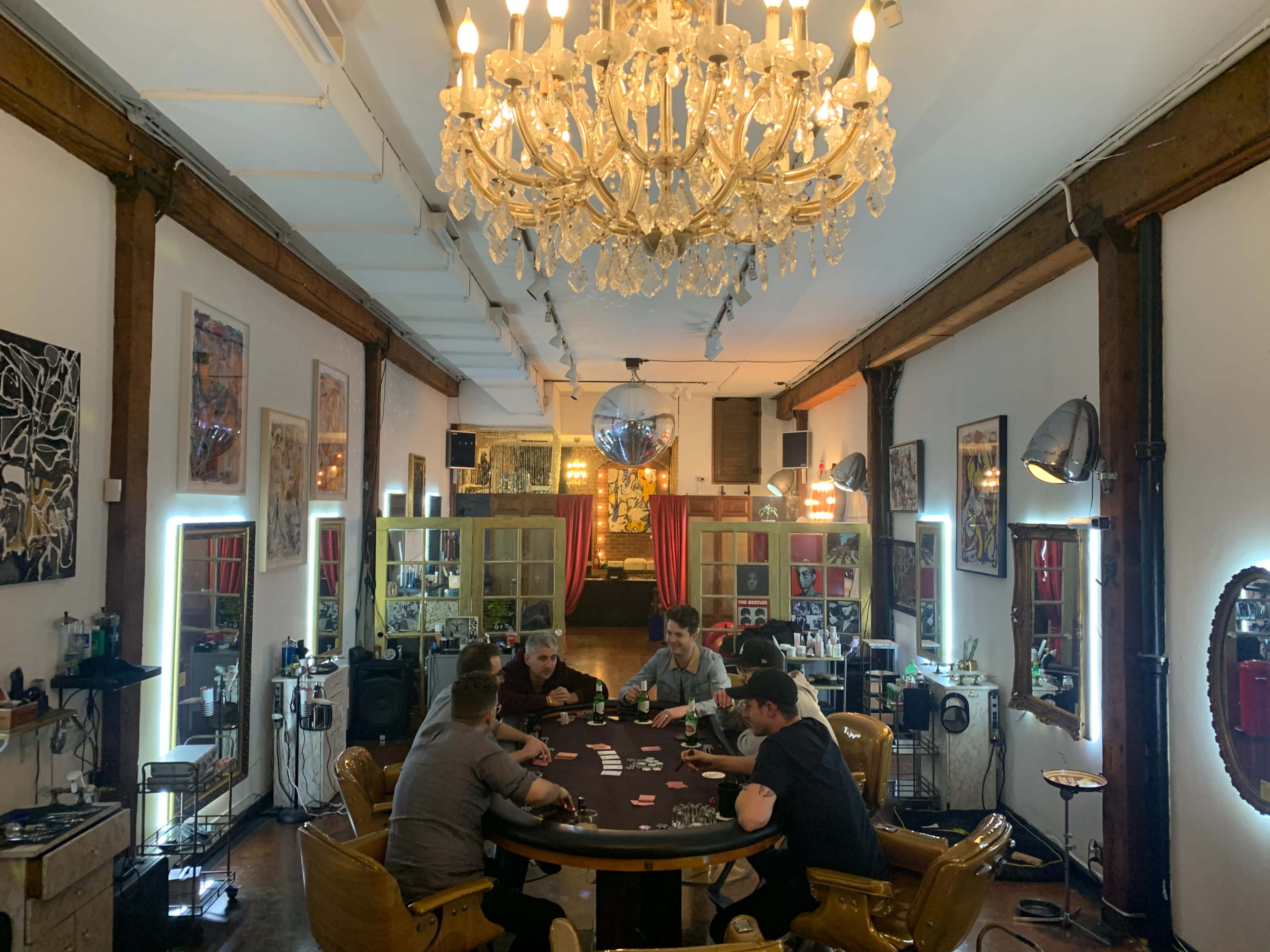 Six people are seated around a circular poker table in a well-decorated room featuring a chandelier and various artworks on the walls.