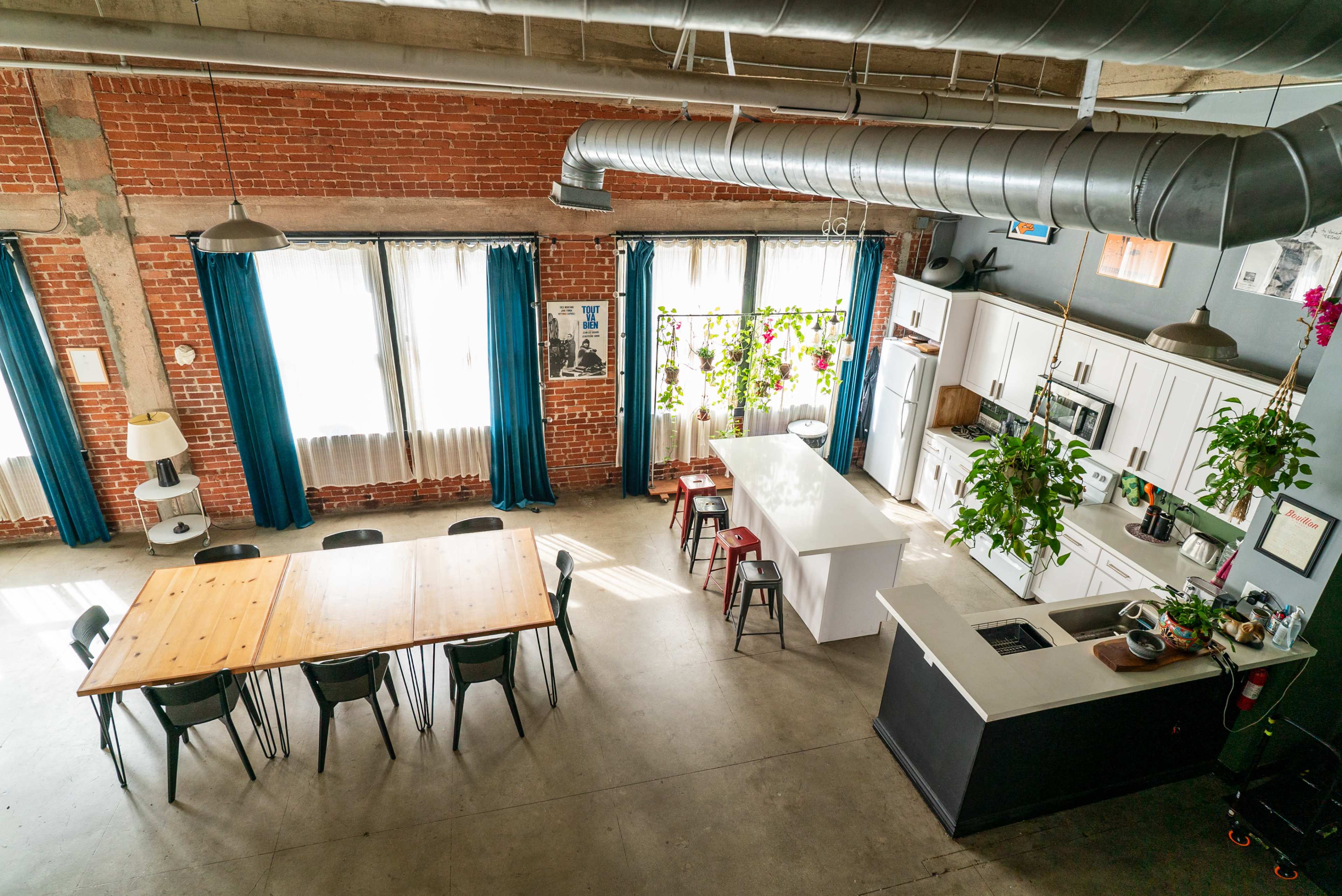 The image depicts a spacious industrial-style kitchen and dining area with exposed brick walls, large windows, and a wooden dining table surrounded by chairs.