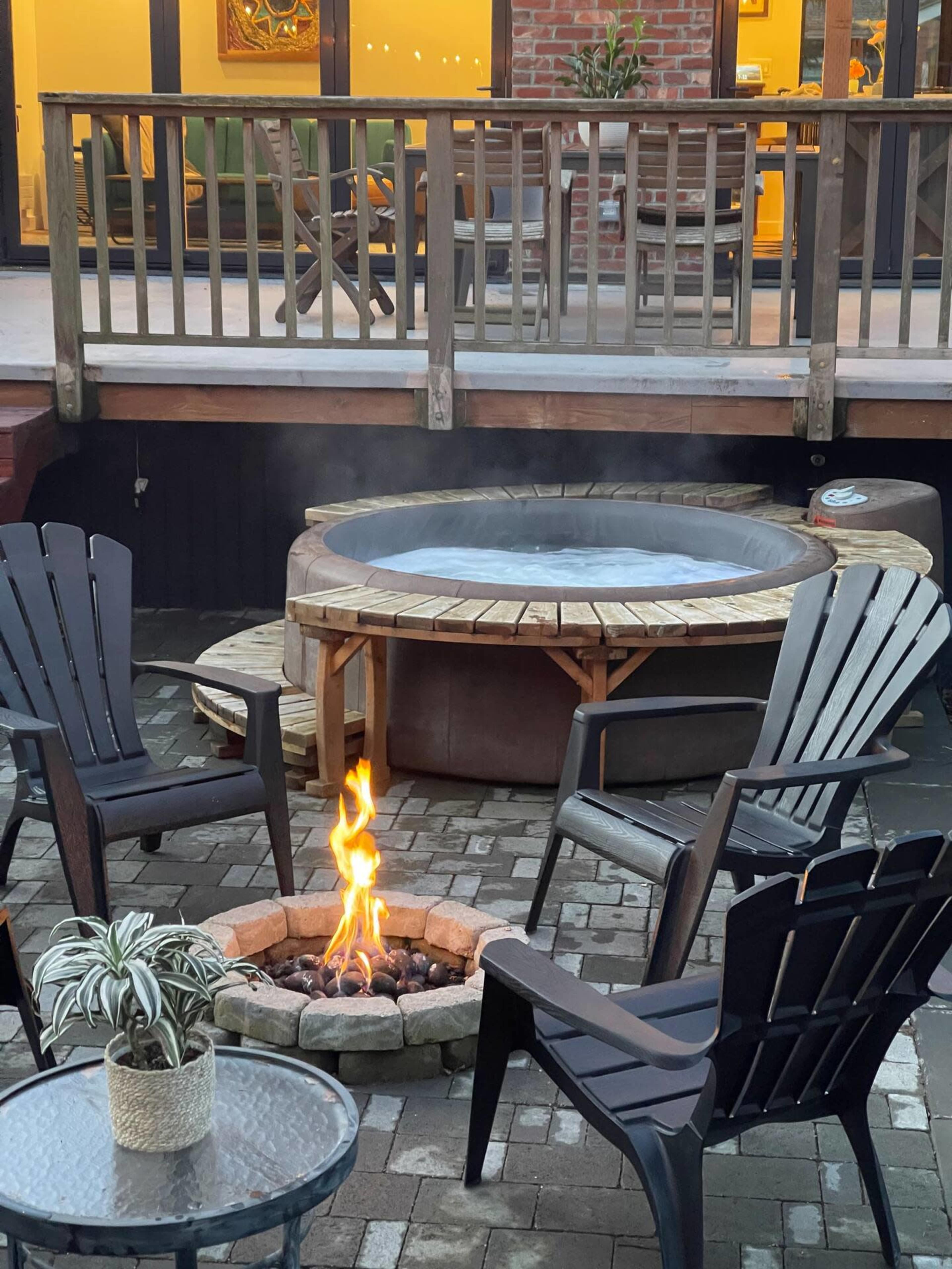 A fire pit with a flame surrounded by black chairs sits next to a hot tub on a patio.