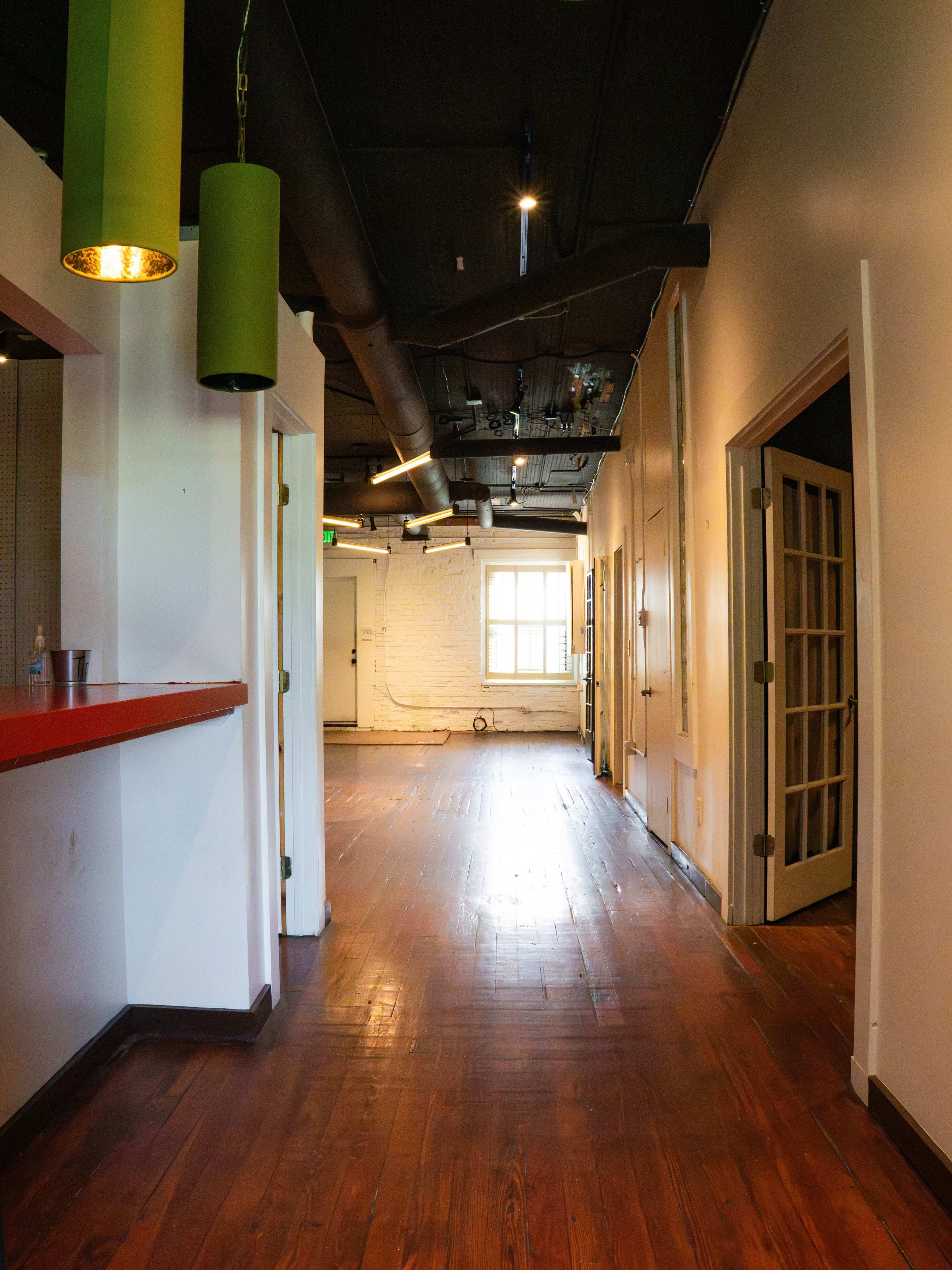 A long, narrow hallway with wooden floors and doors leading to adjacent rooms, illuminated by overhead lighting and green pendant lamps.
