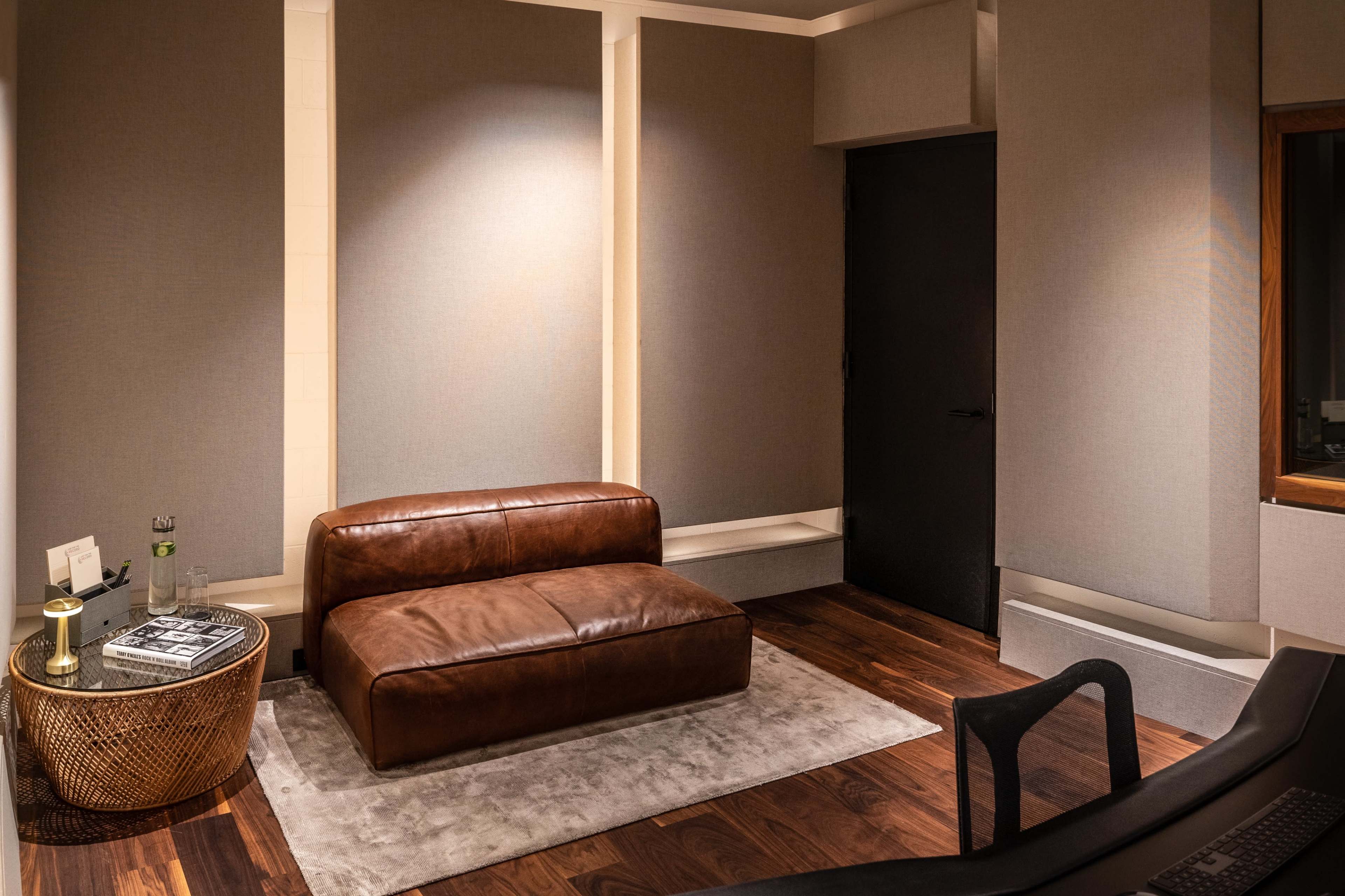 The image shows a modern interior featuring a brown leather couch, a round side table, and a dark door, with soundproofed walls and wooden flooring.