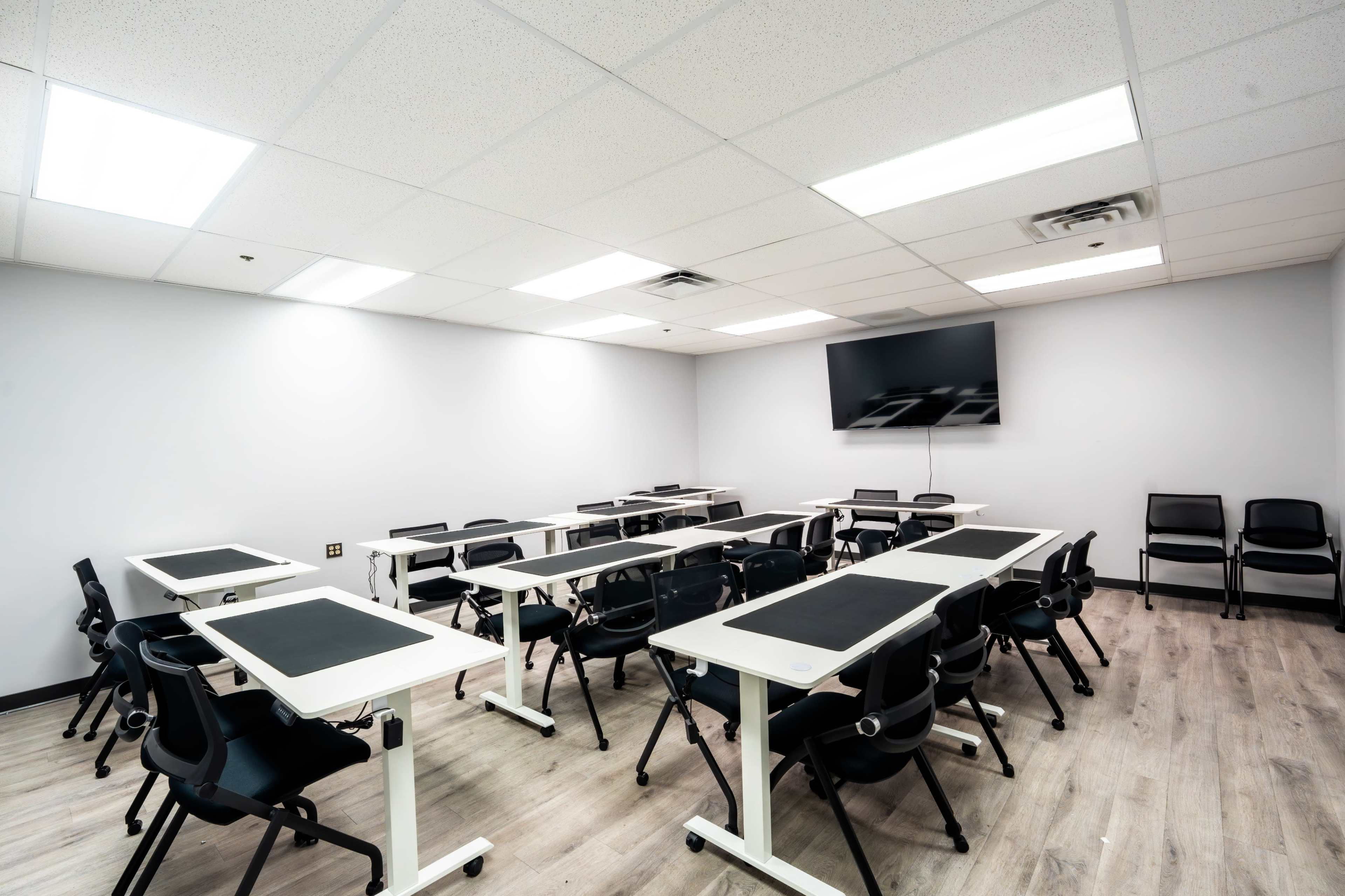 The image shows a modern classroom setup with multiple desks and chairs arranged in a U-shape, and a large screen mounted on the wall.