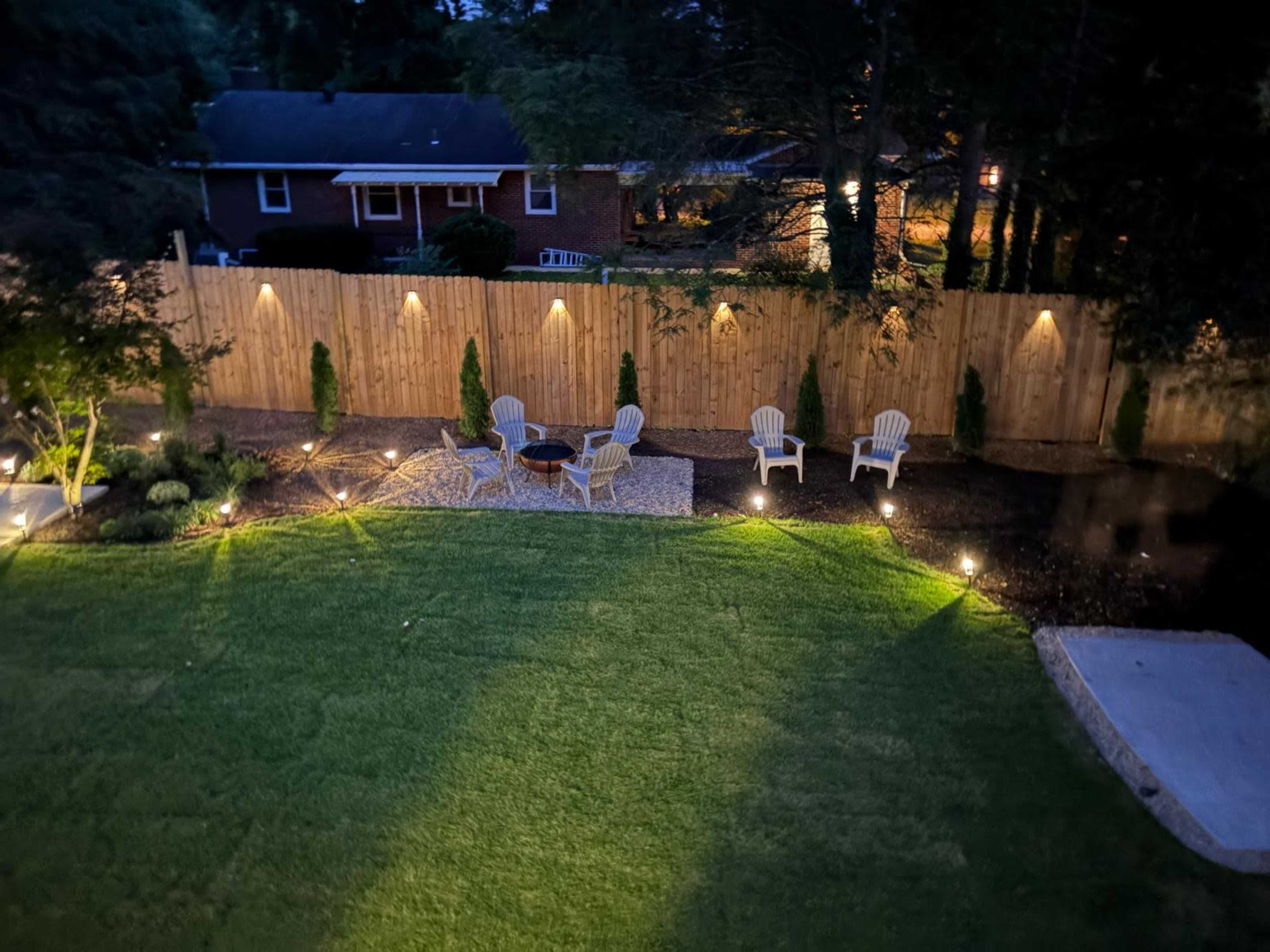 The image shows a well-lit backyard with four white chairs arranged around a circular stone area, bordered by a wooden fence and trees.