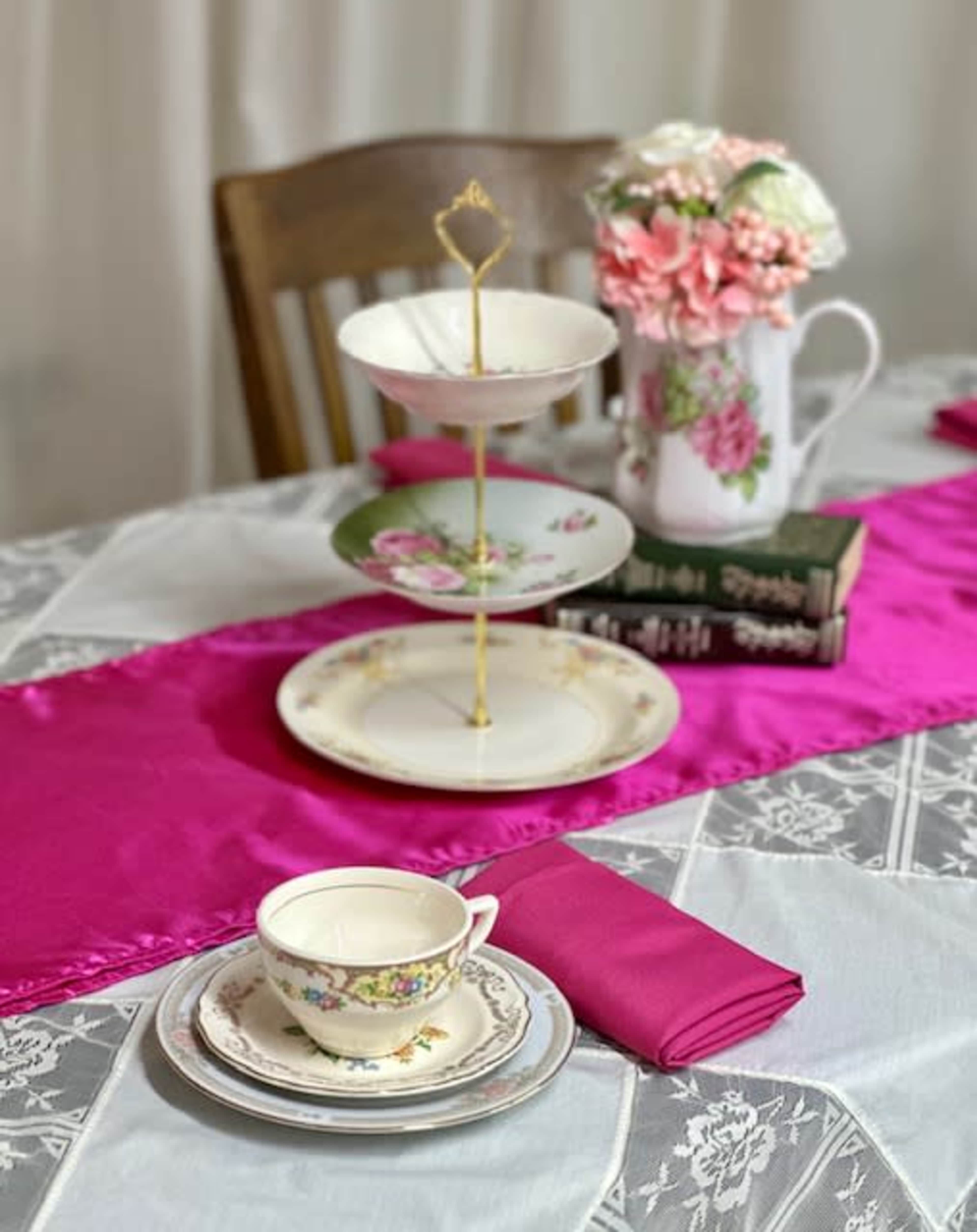 A tiered serving stand is set on a table with a pink table runner, along with a delicate teacup, floral pitcher, and stacked books.