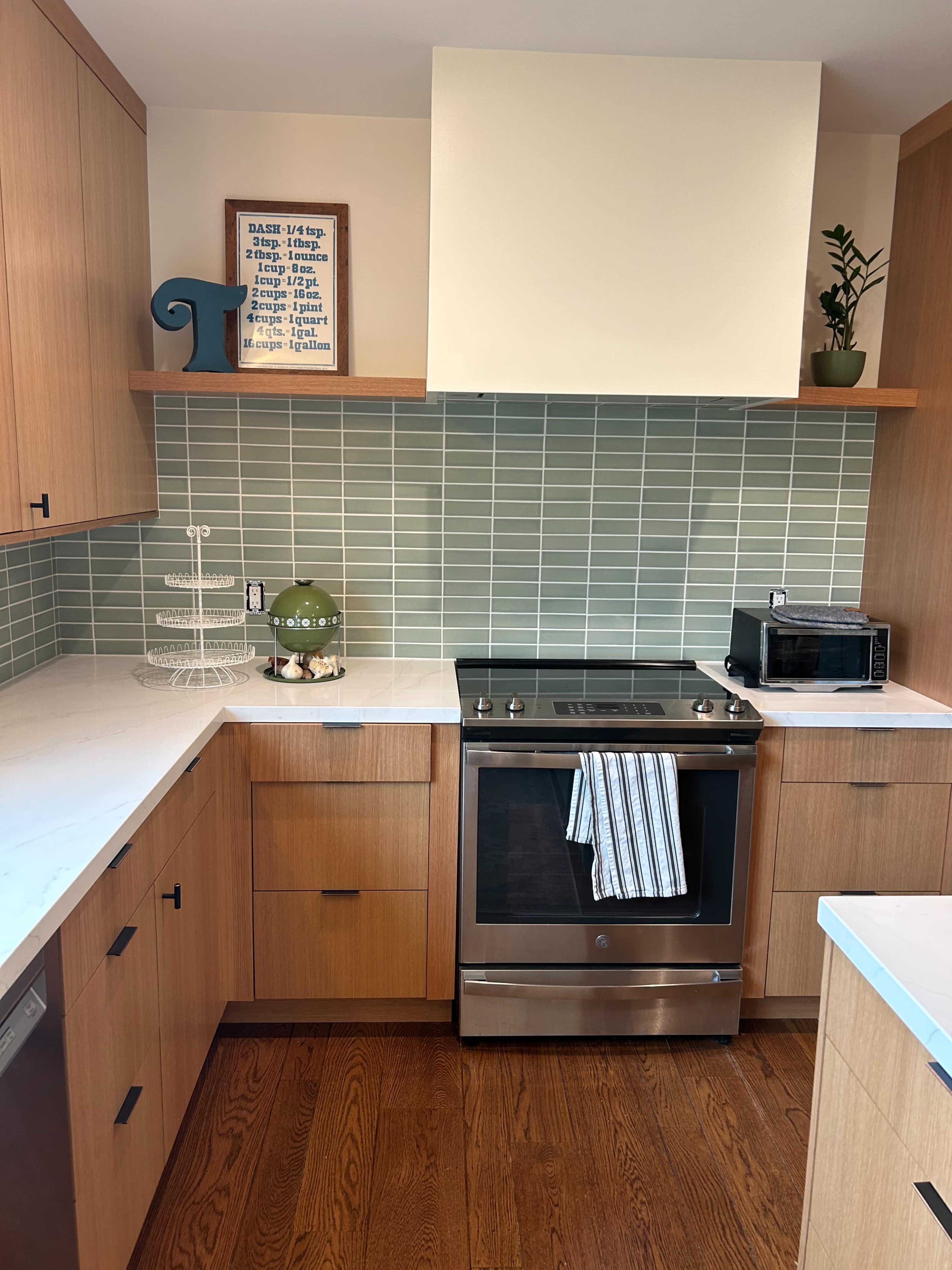 The image shows a modern kitchen featuring wooden cabinets, a stainless steel oven, and a tiled backsplash in muted green shades.