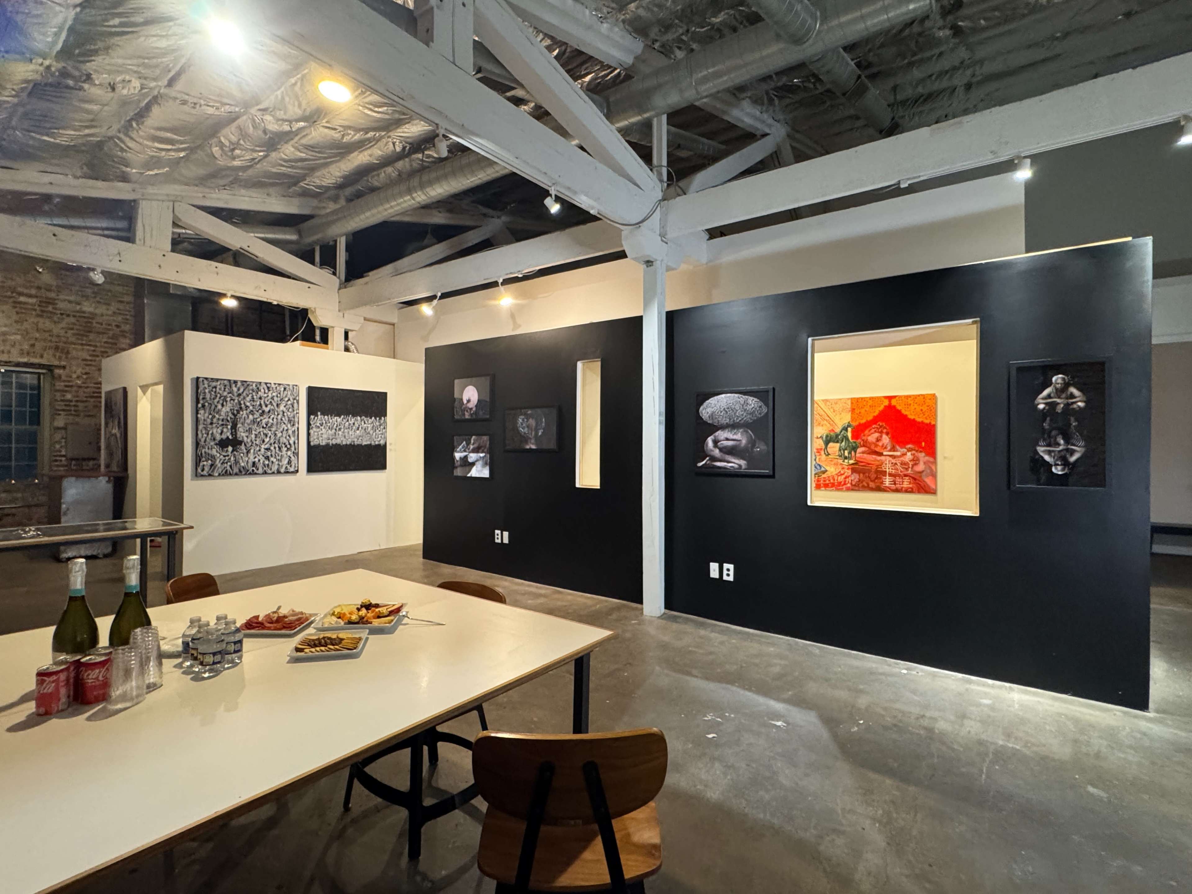 The image shows an art gallery featuring walls with various framed artworks, a table with food and drinks, and exposed beams on the ceiling.