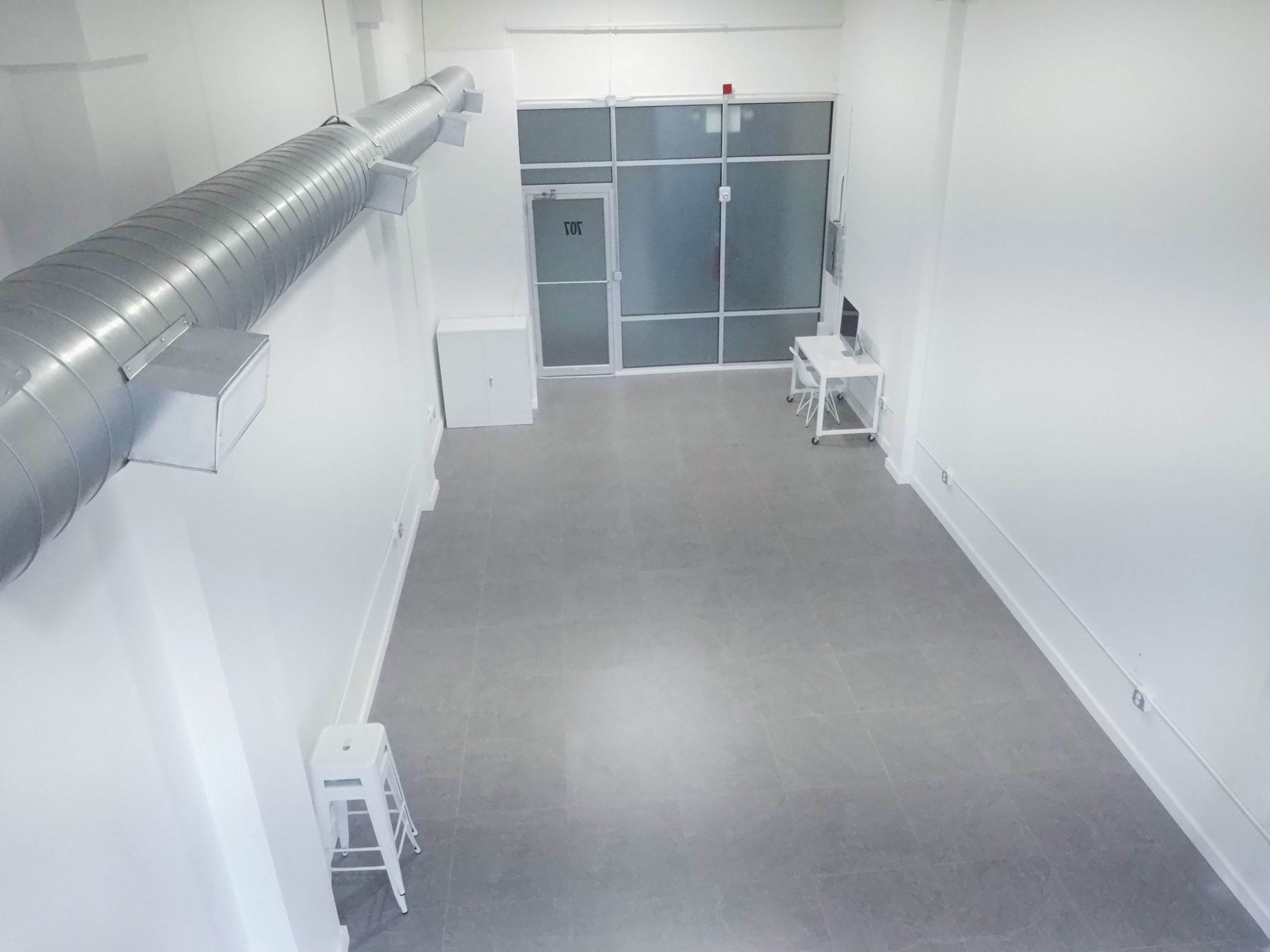 A large, empty room with grey tiled flooring and white walls, featuring a metallic duct overhead and a glass partition at one end.
