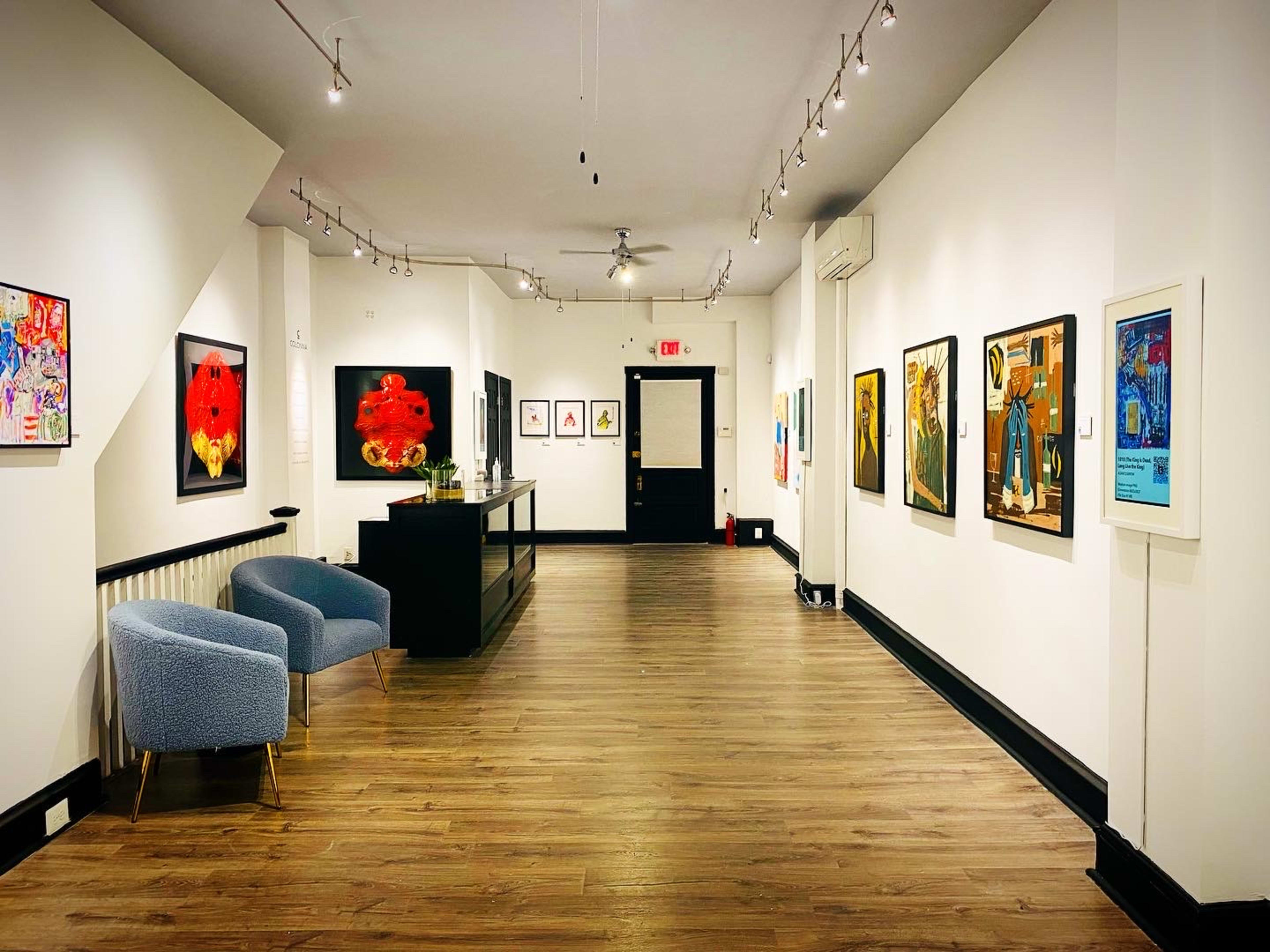 An art gallery with several paintings displayed on white walls, illuminated by track lighting, and featuring a blue chair and a reception desk.