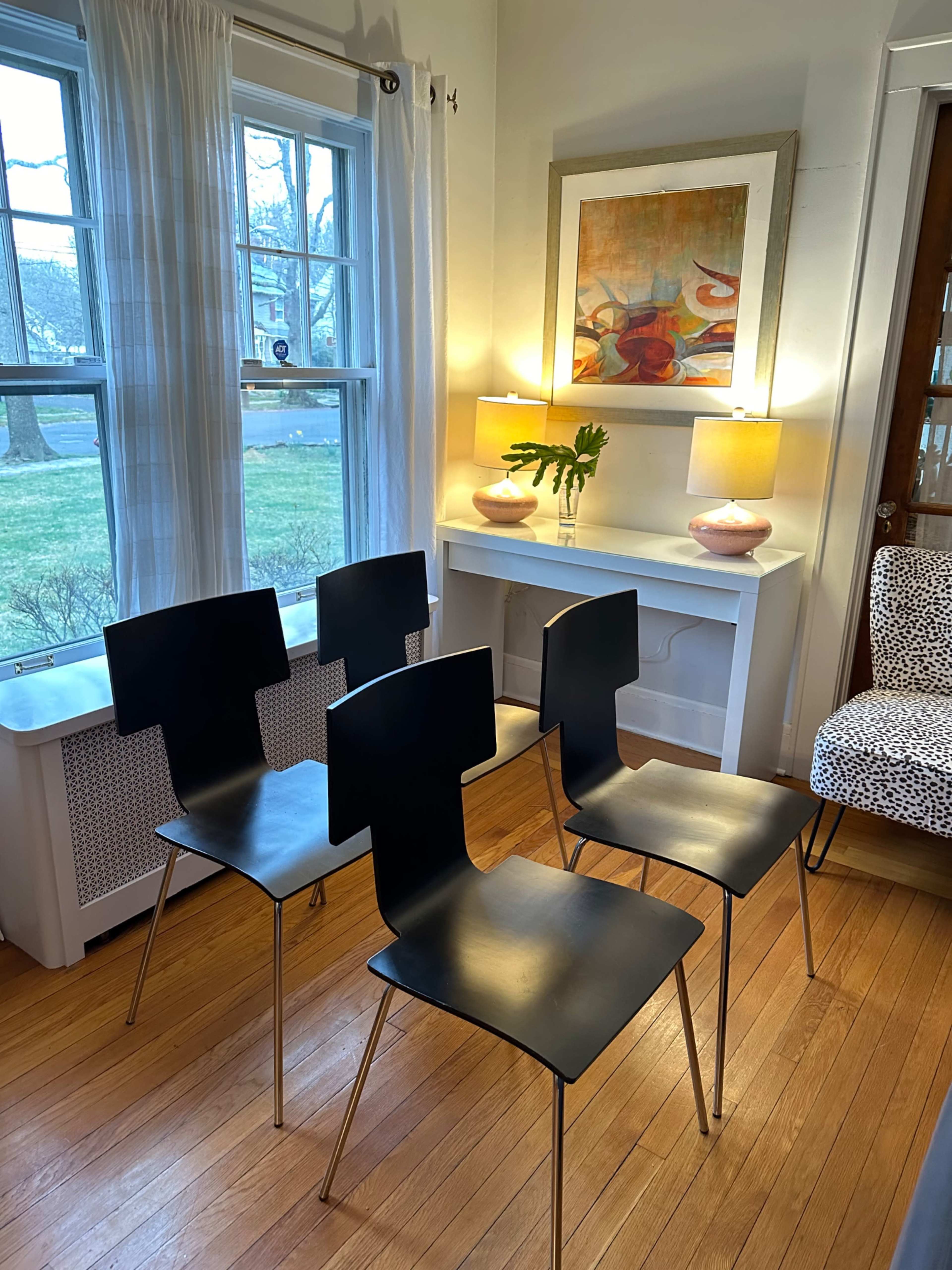 Four black chairs with unique backs are arranged in a bright room featuring windows, a small table with lamps, and a colorful piece of artwork on the wall.