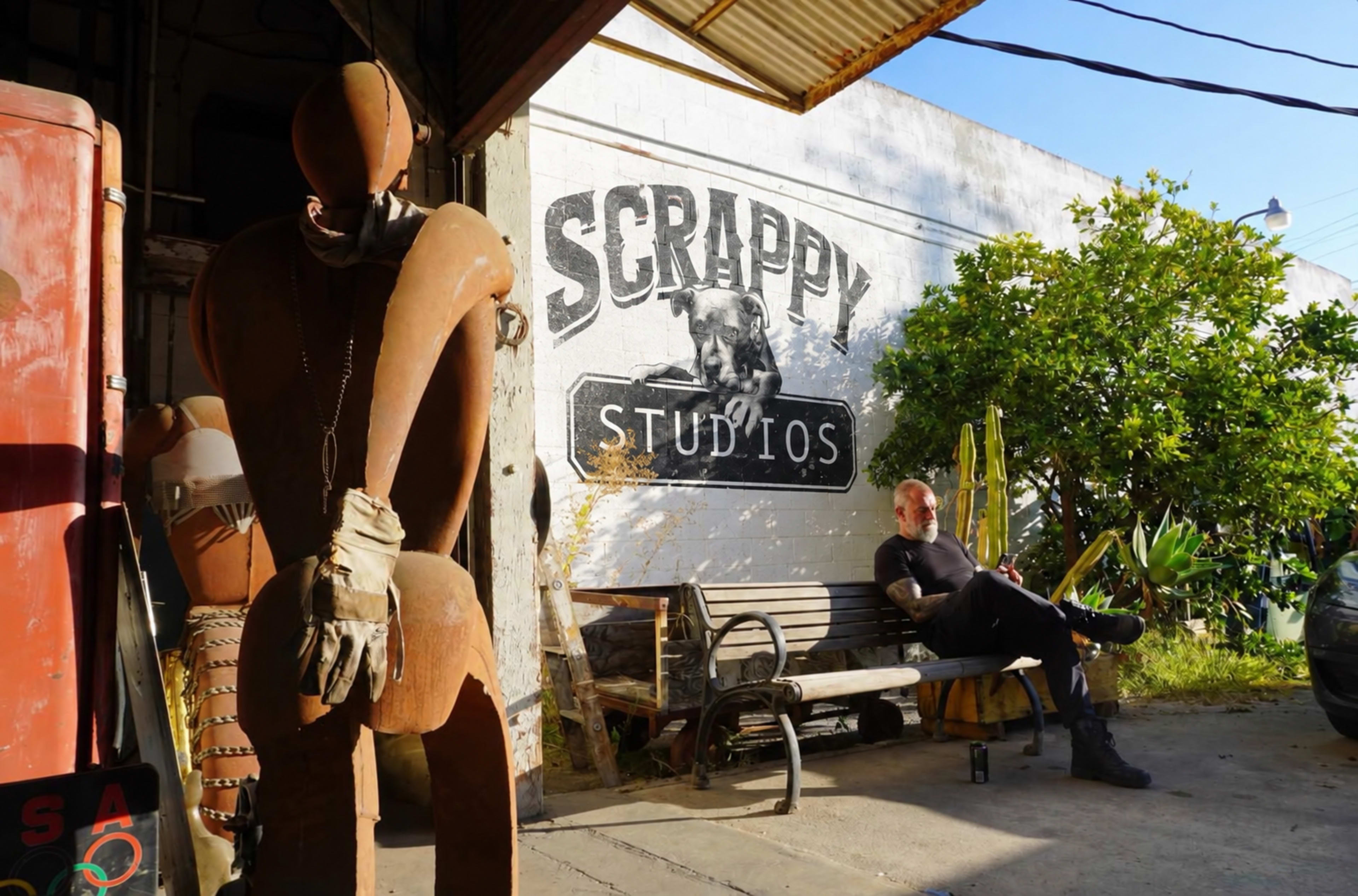 A rusted metal sculpture stands beside a man sitting on a bench in front of a studio with a painted sign reading "SCRAPPY STUDIOS."