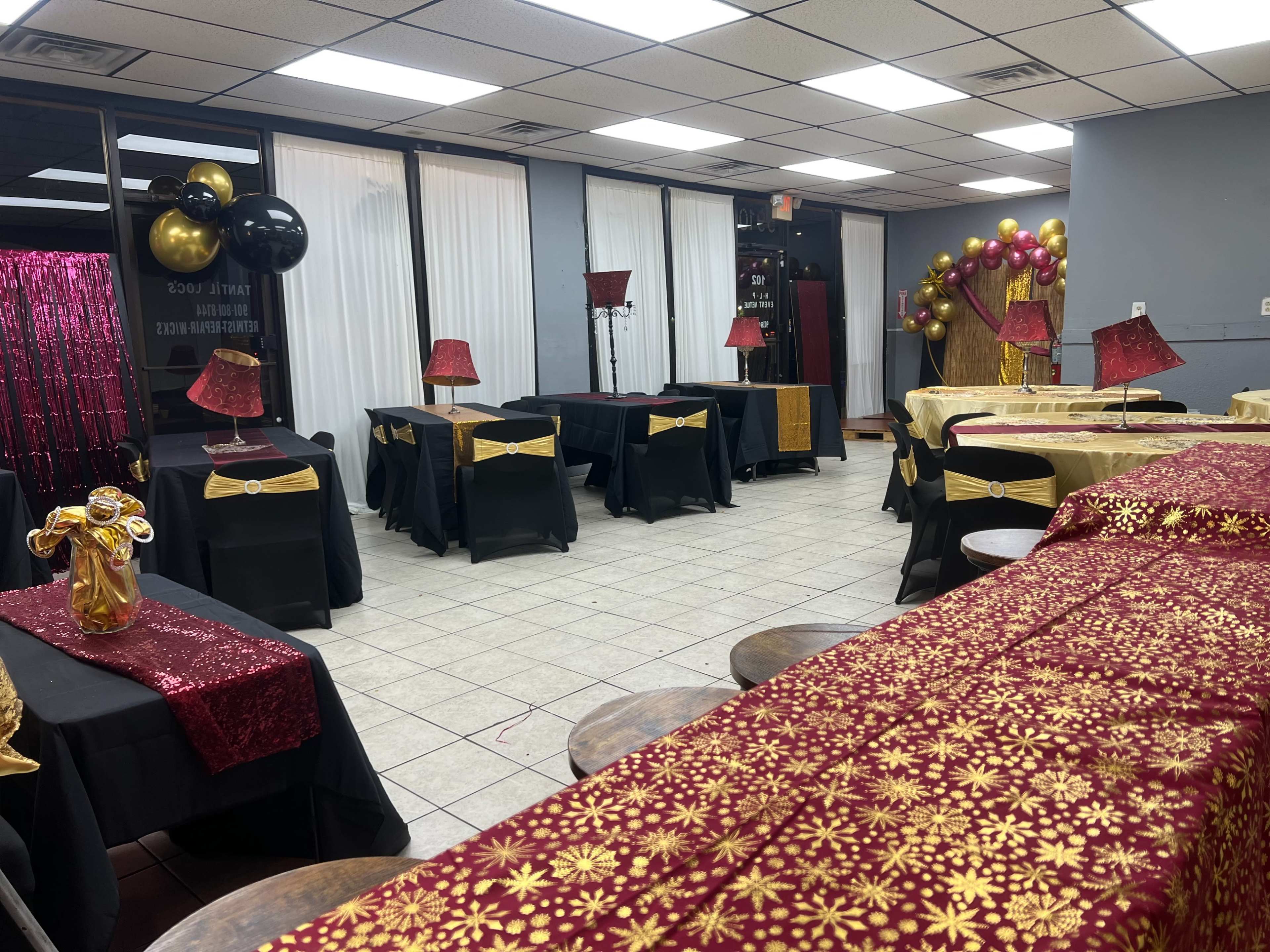 The image shows a decorated event space with several tables covered in black and gold tablecloths, illuminated by lamps, and featuring balloons and a sparkling backdrop.