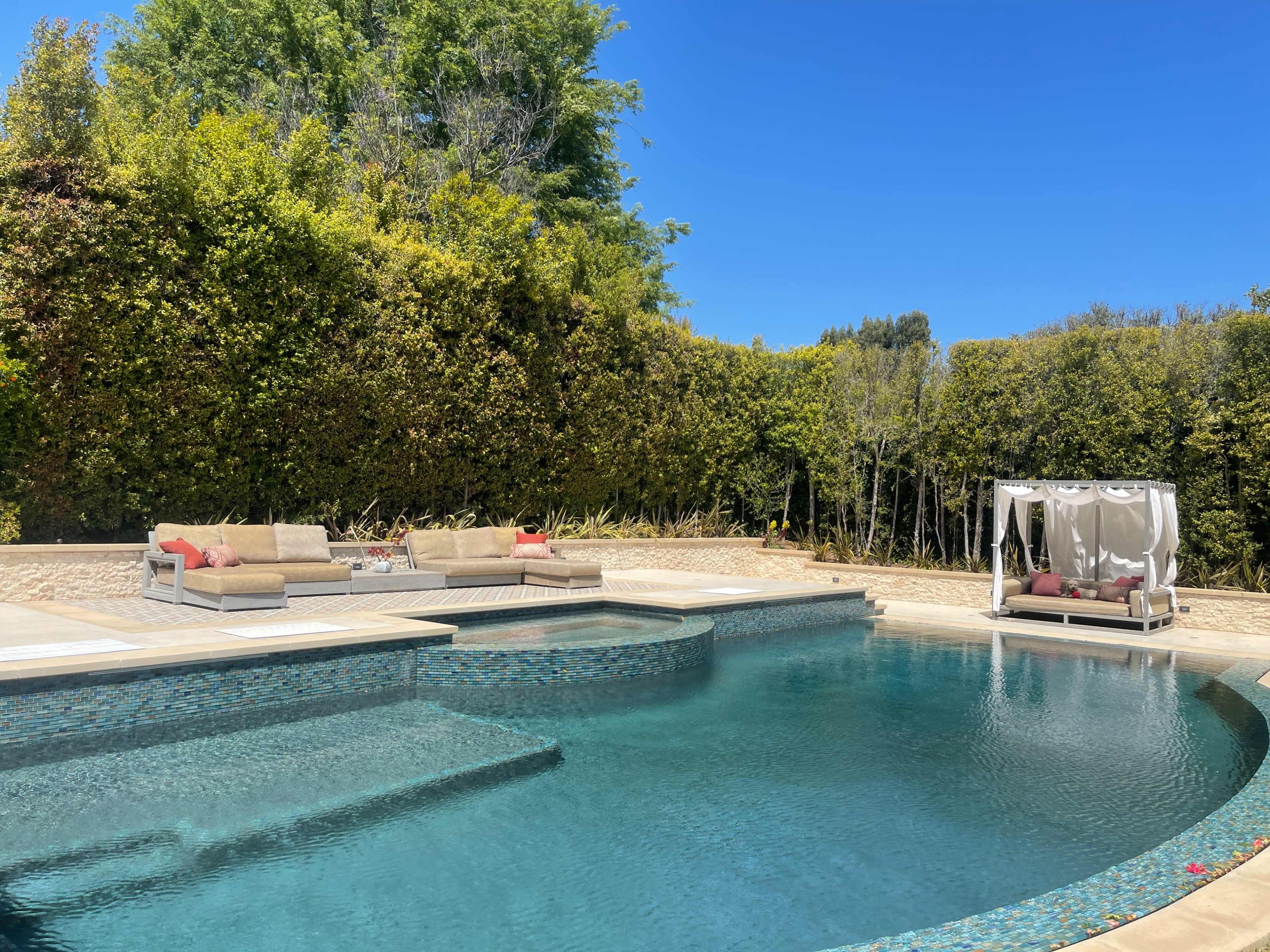 A spacious outdoor pool is surrounded by comfortable seating and a shaded lounge area, with lush greenery in the background.