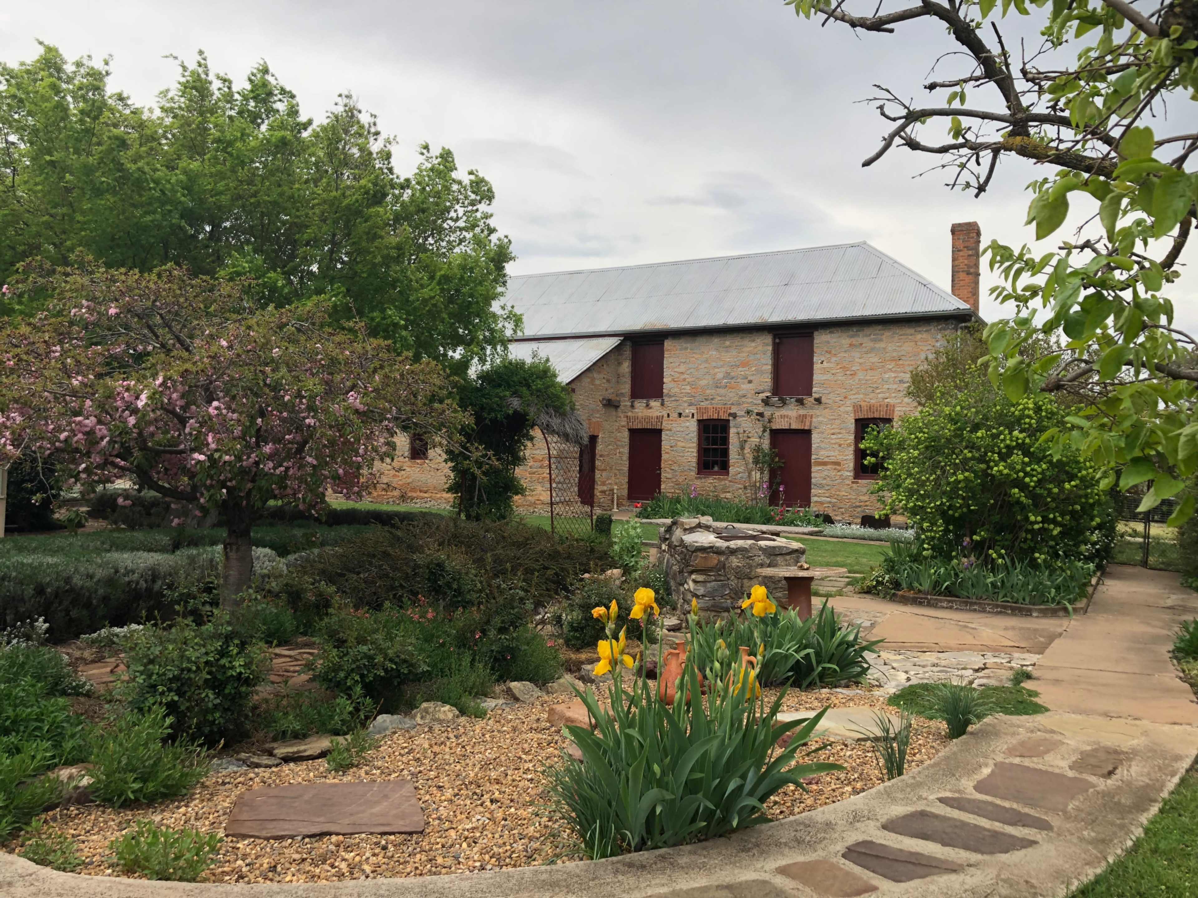 A brick house with a metal roof stands behind a landscaped garden featuring flowers and trees.