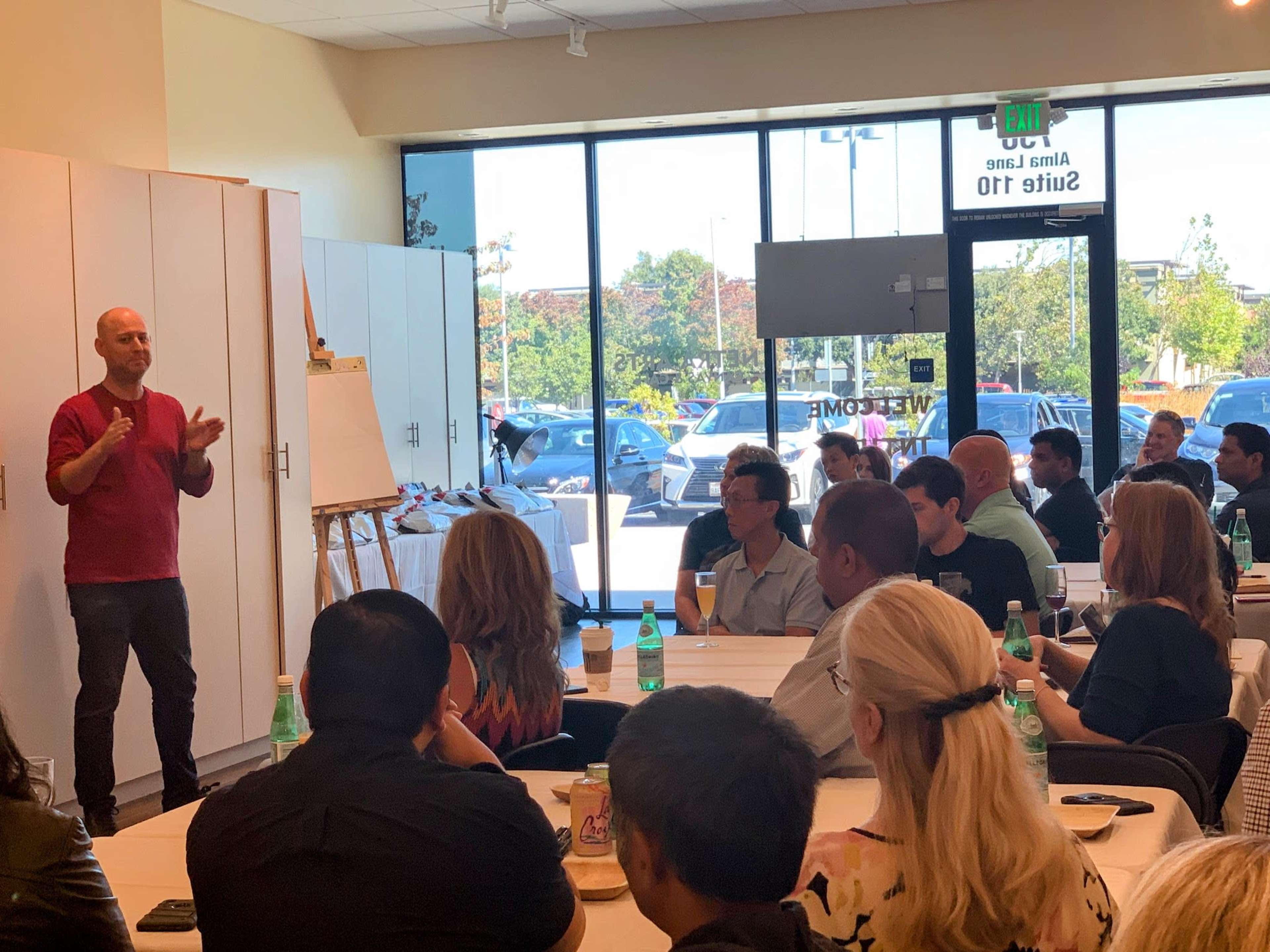 A person stands at the front of a room presenting to an audience seated at tables.